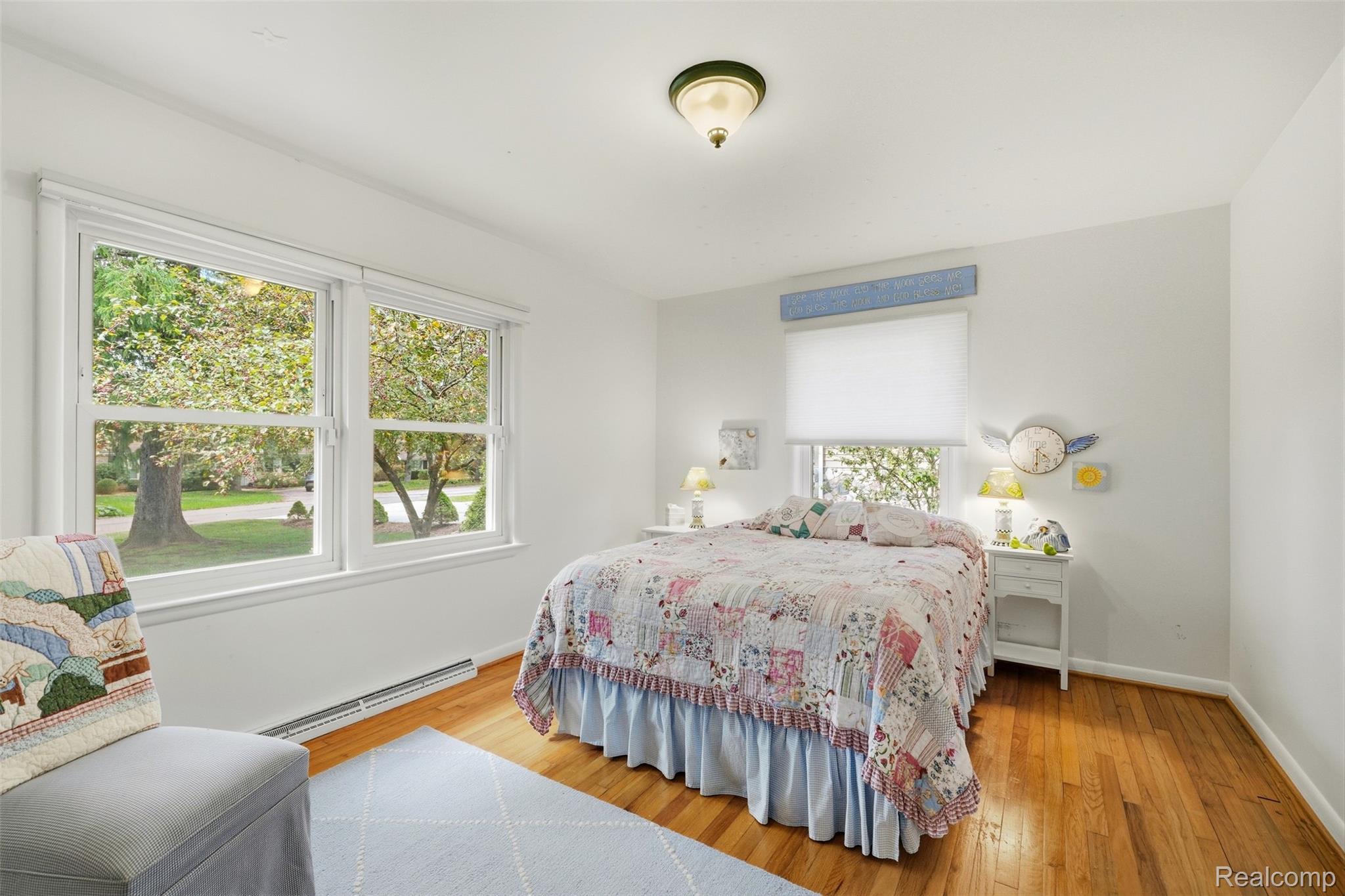 Bedroom with a floral patchwork bedspread, hardwood floors, and large windows overlooking trees.