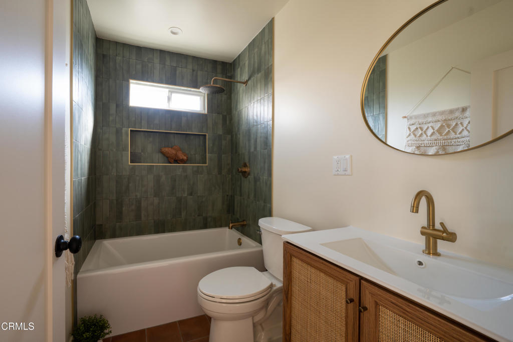 Bathroom with a green tiled tub and shower, woven-front vanity, and brass fixtures.