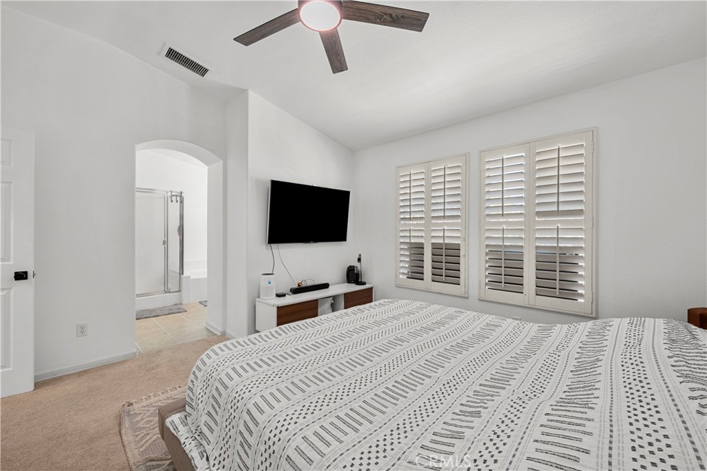 Bedroom with plantation shutters, a patterned bedspread, and an arched doorway to a bathroom.