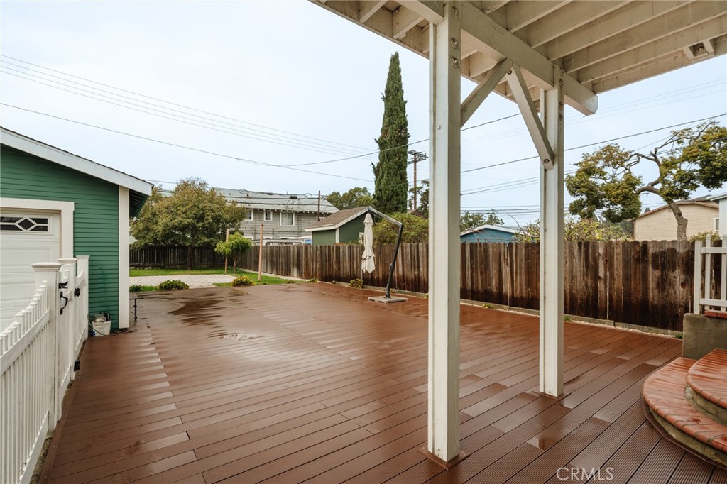 Backyard deck beneath a white pergola, with a fenced yard and green outbuilding.