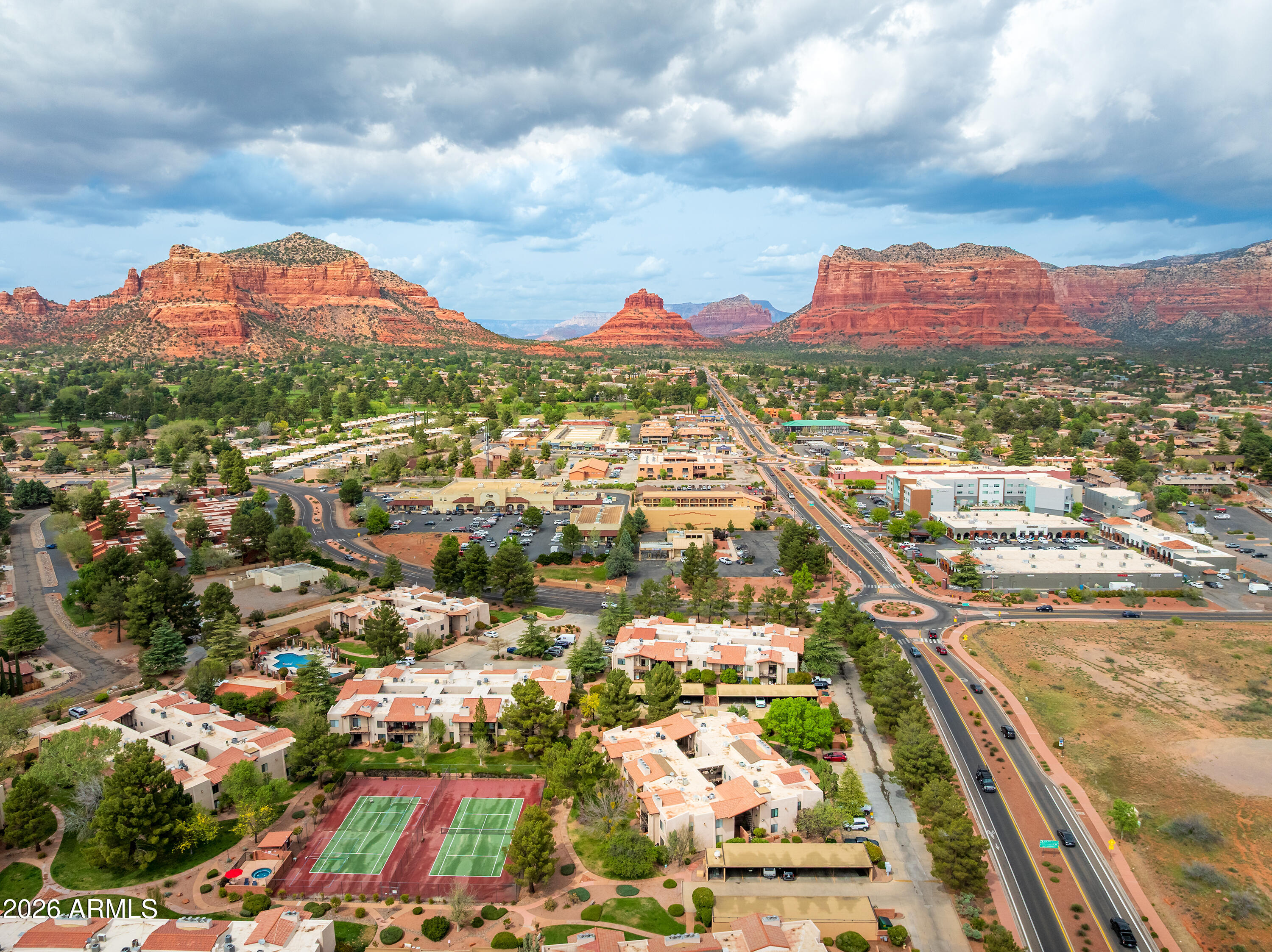 Aerial view of a Sedona neighborhood beneath red rock formations, with tennis courts and a main road.