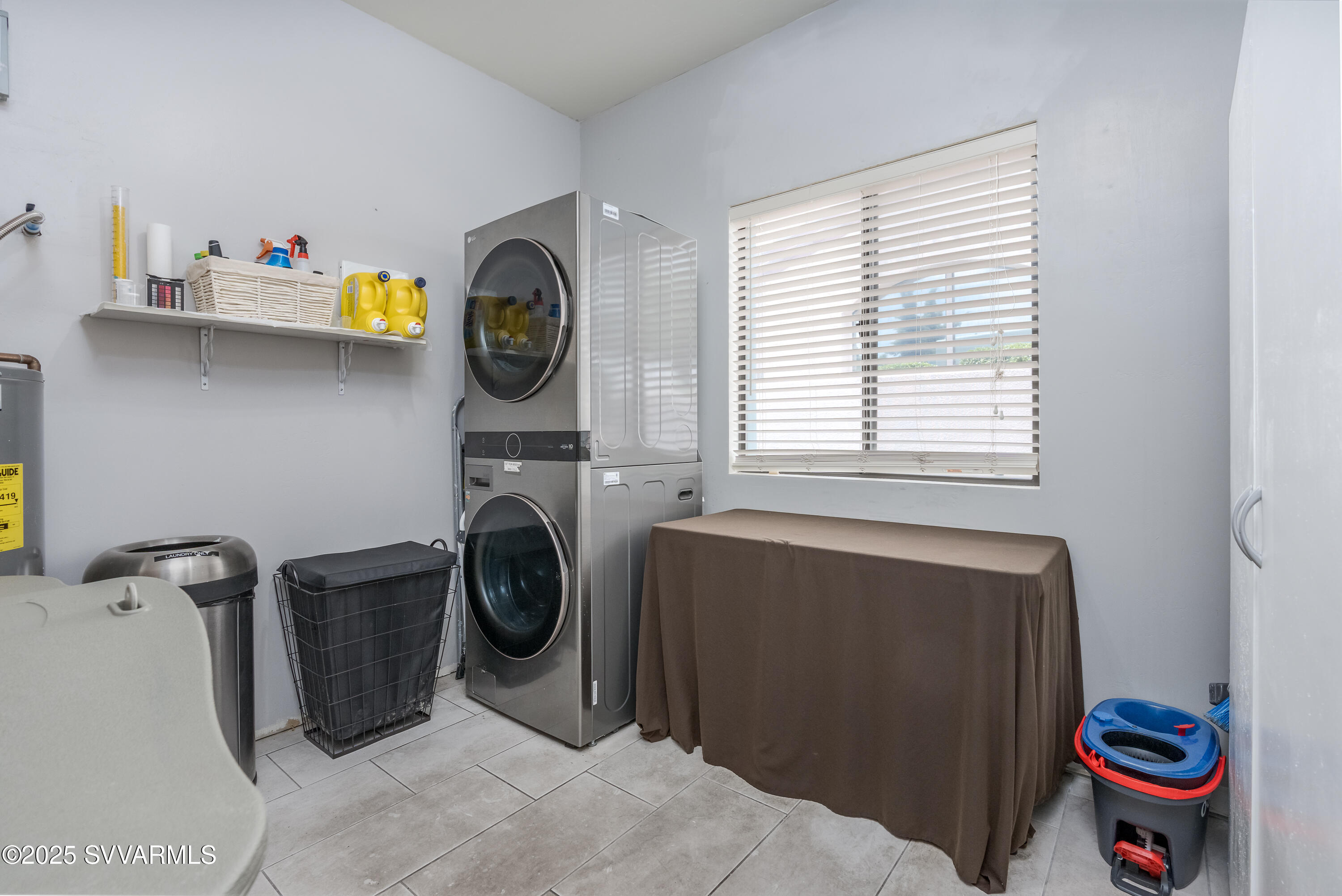 Laundry room with stacked stainless steel washer and dryer, cleaning supplies, and a covered table.