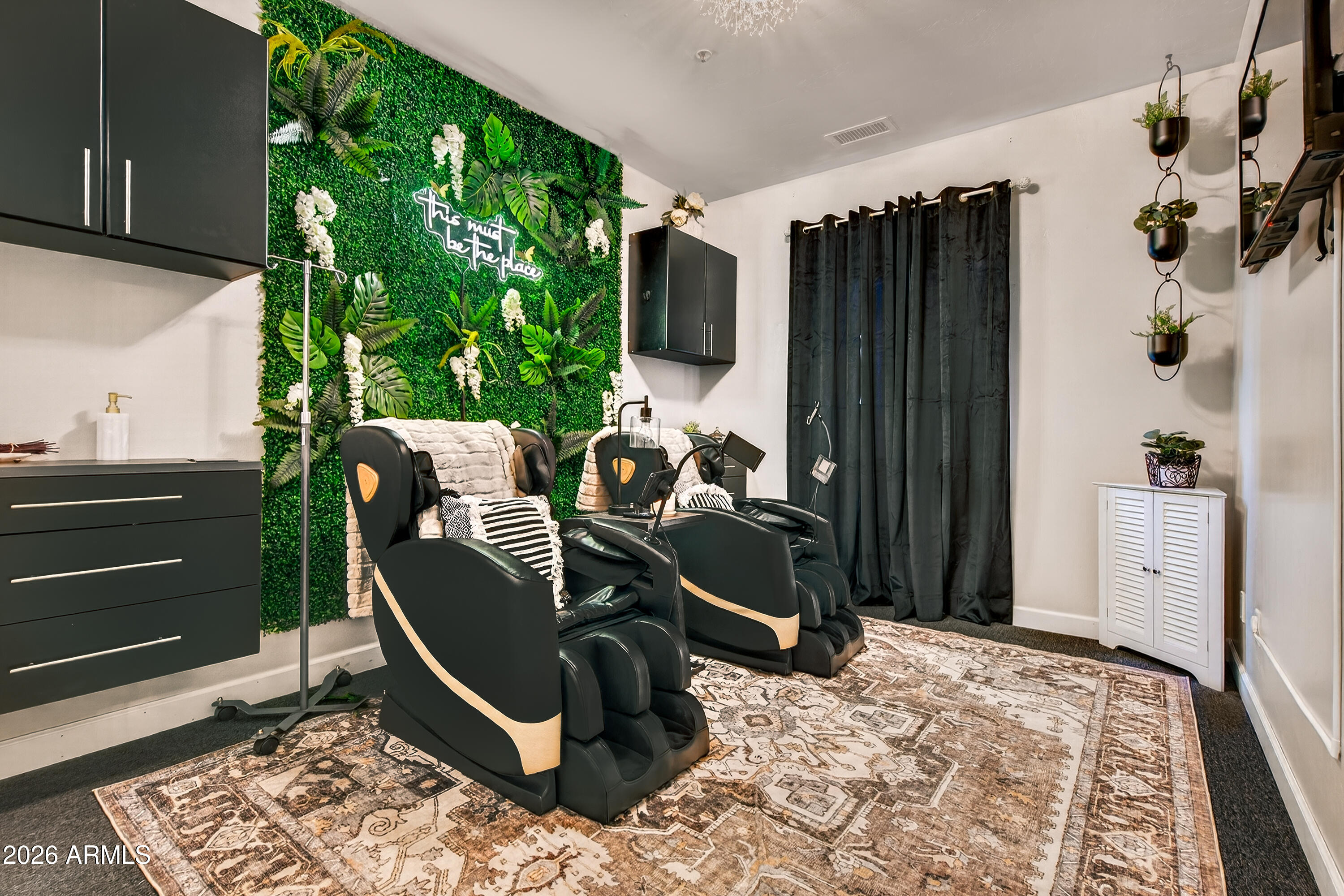 Furnished room with two massage chairs, a leafy feature wall, dark curtains, and a patterned rug.