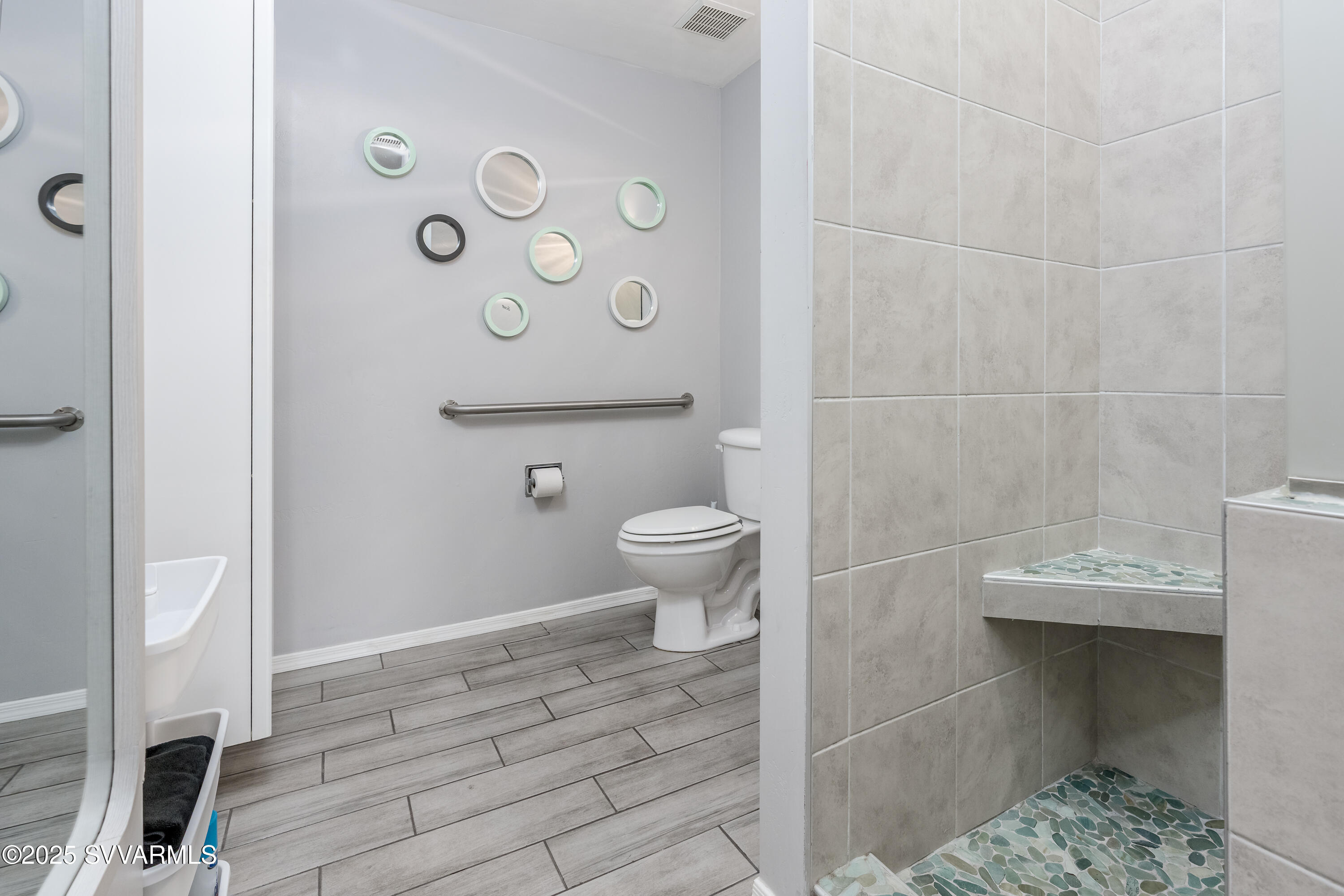 Bathroom with a pebble-mosaic walk-in shower, built-in bench, toilet, grab bar, and round mirrors.