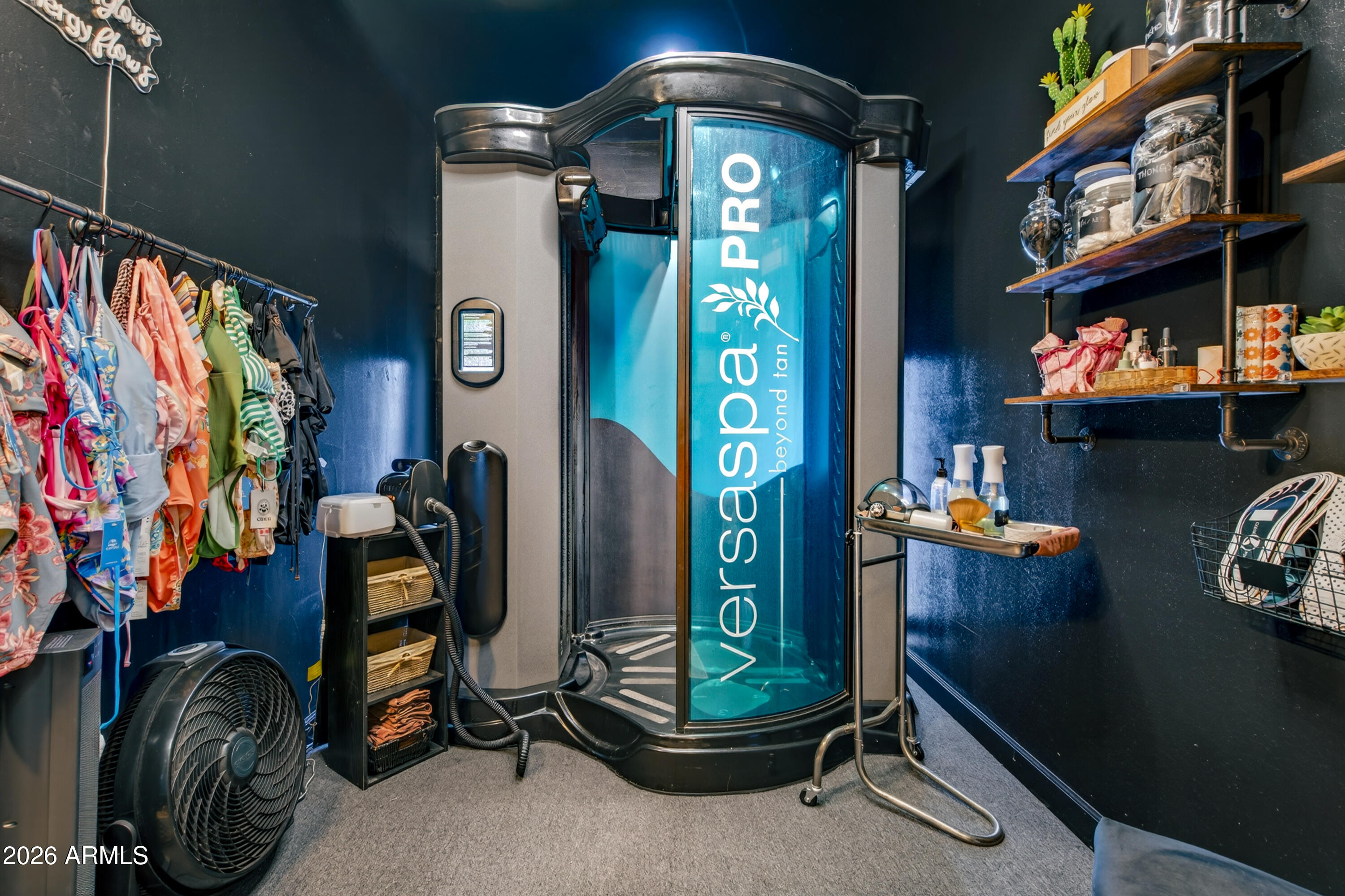 Interior room with a VersaPro tanning booth, clothing rack, supply shelves, and floor fan.