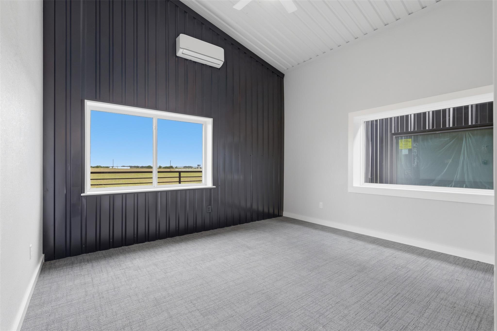 Vacant carpeted room with a dark paneled wall, mini-split air conditioner, and window overlooking a field.