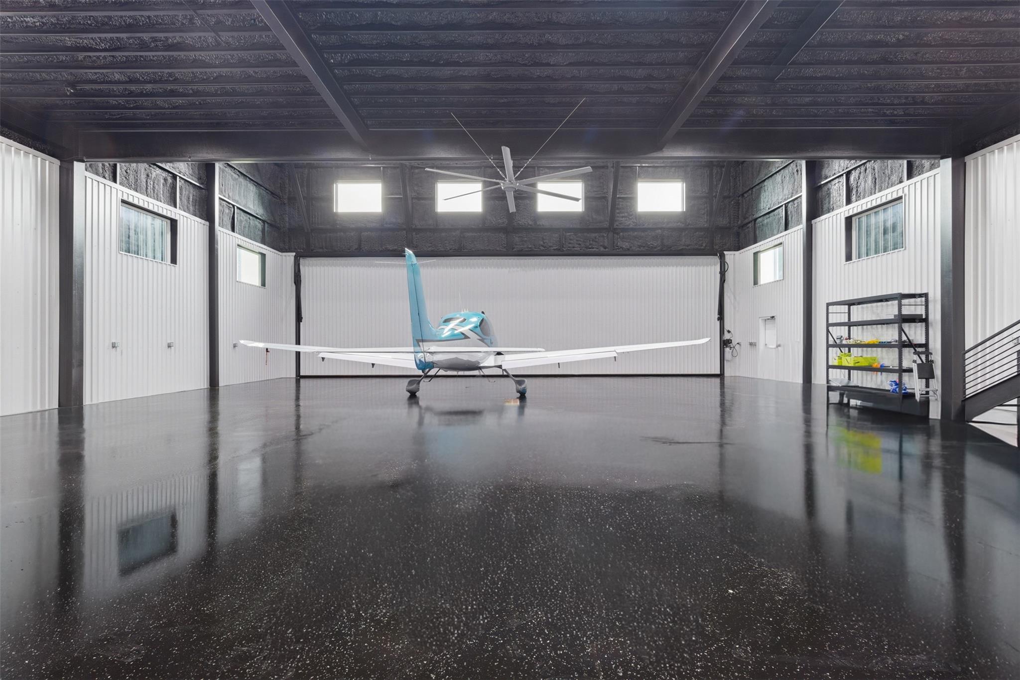 Aircraft hangar with a blue-and-white plane, glossy floor, high ceiling, and large fan.