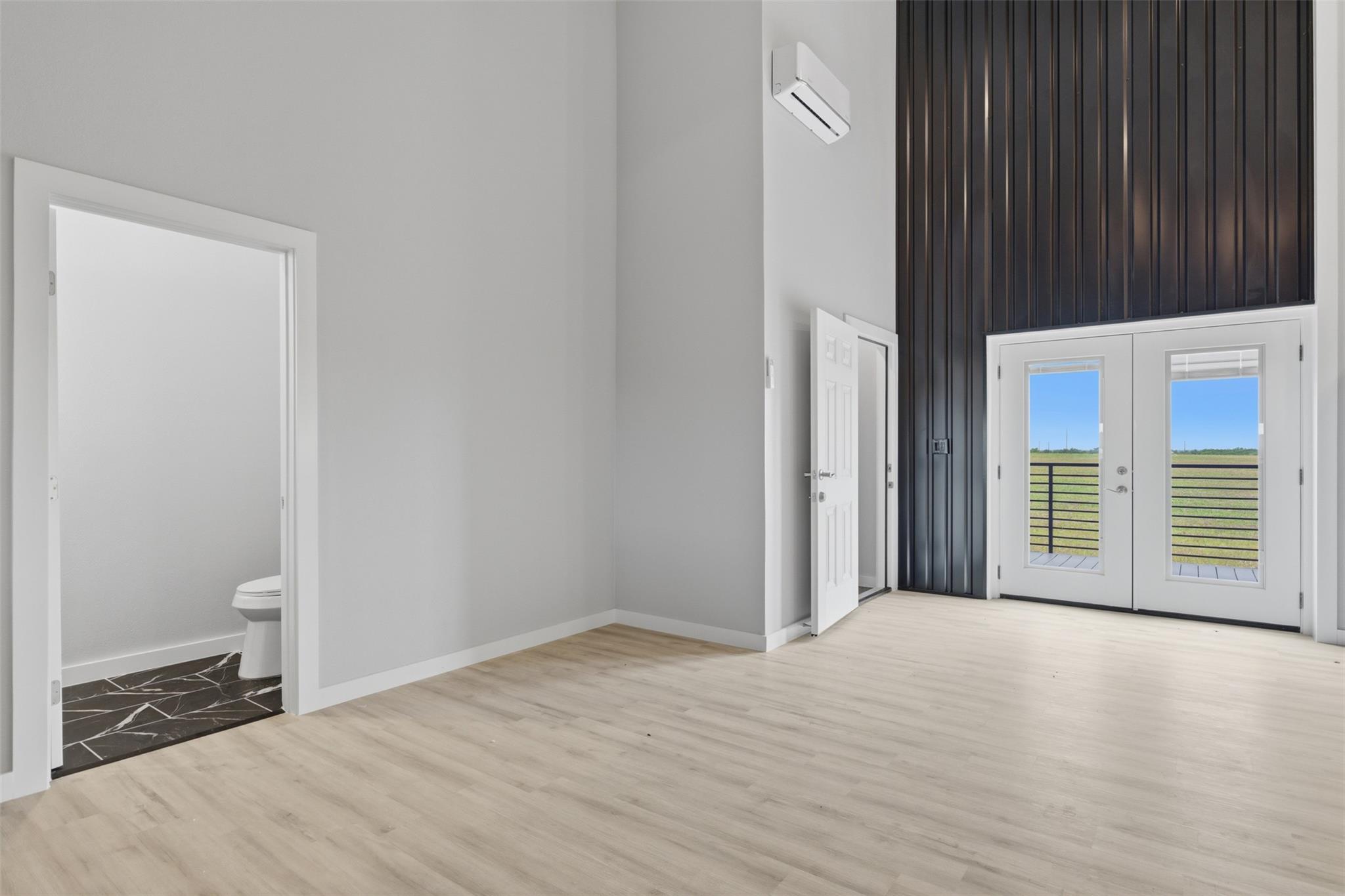 Vacant room with pale wood-look floors, dark paneled wall, and double doors overlooking a grassy field.