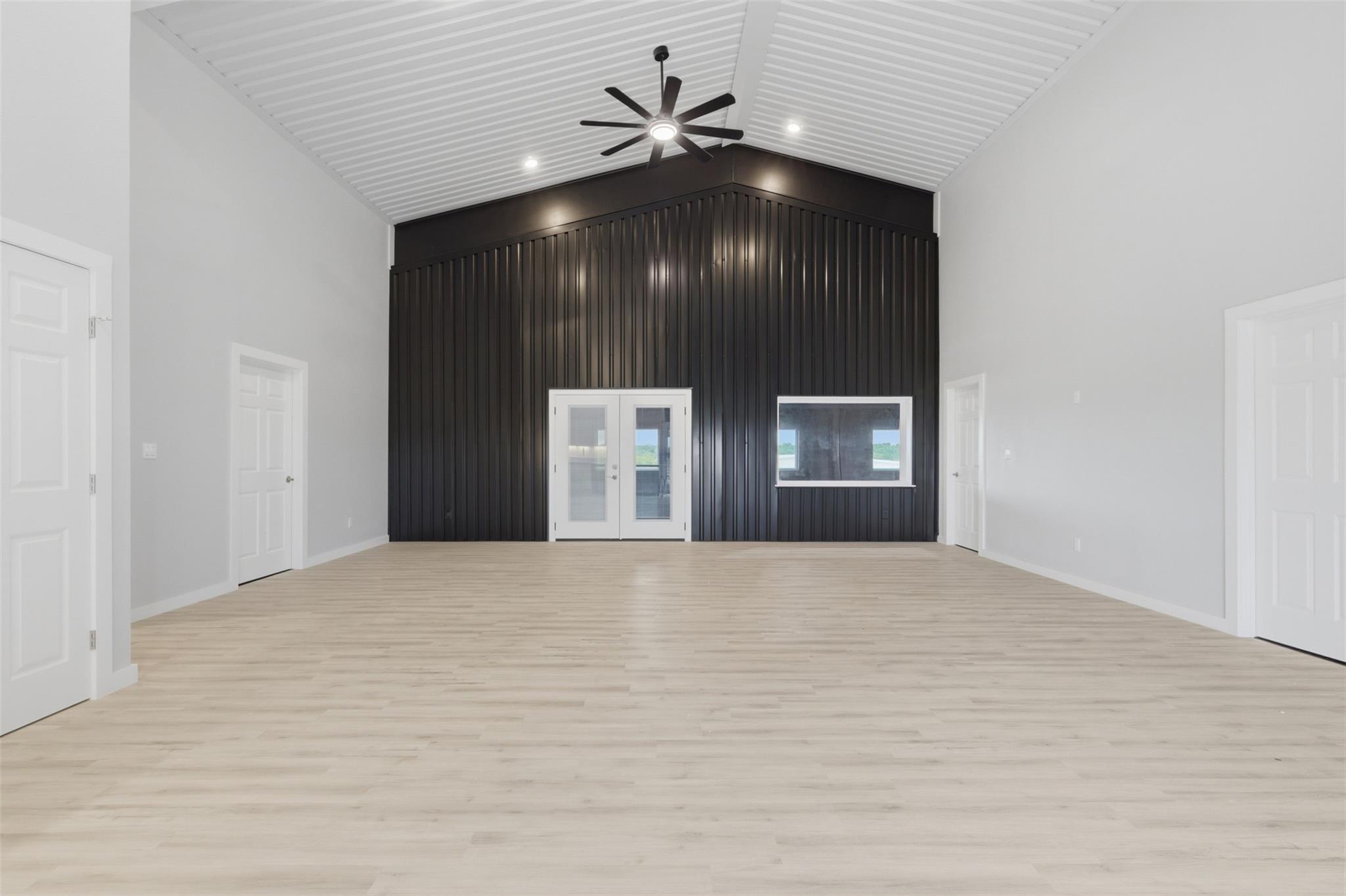 Empty vaulted room with pale wood-look floors, a dark paneled wall, and glass double doors.