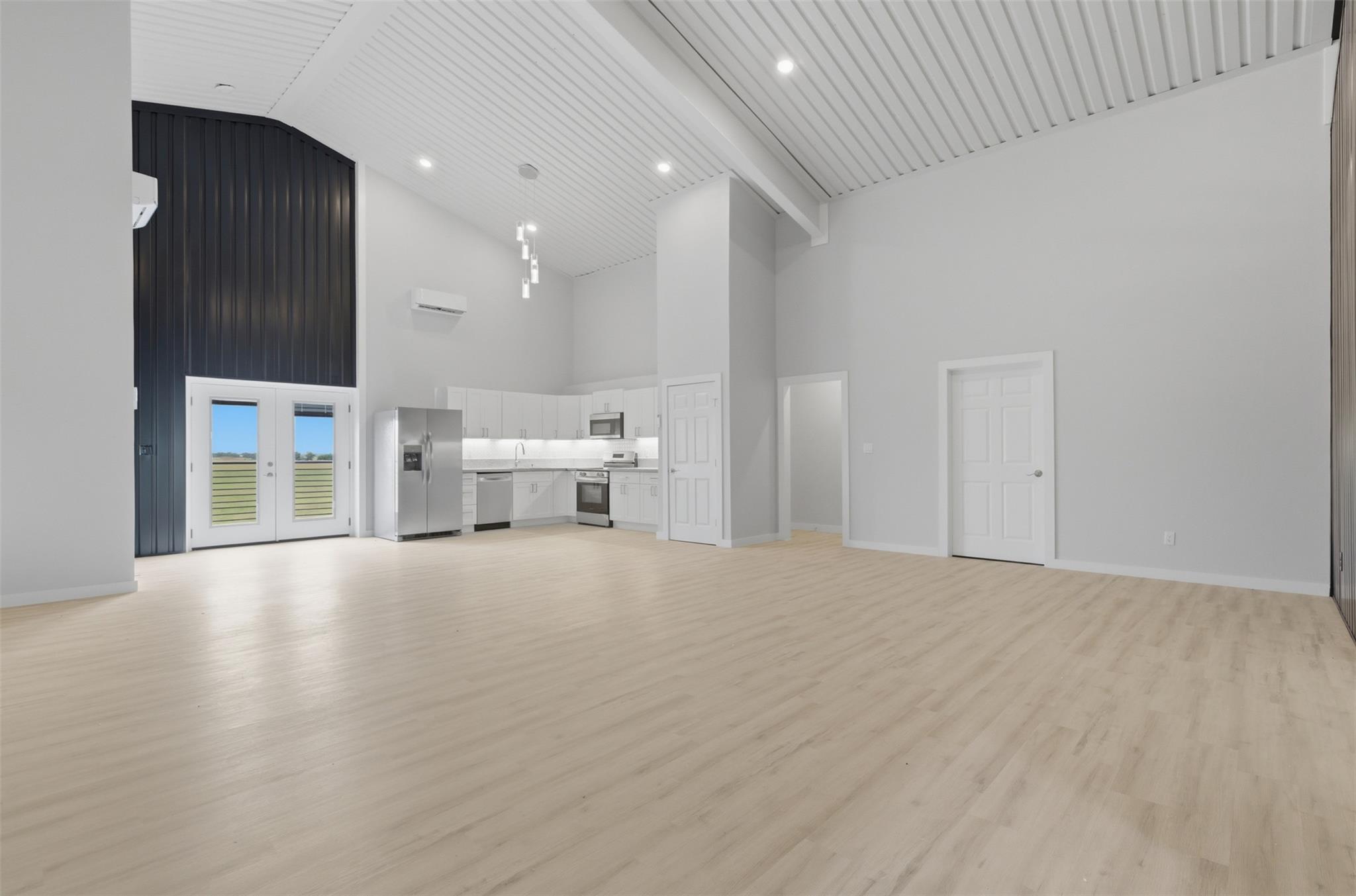 Open-plan kitchen and living area with vaulted ceiling, pale wood floors, and glass double doors.