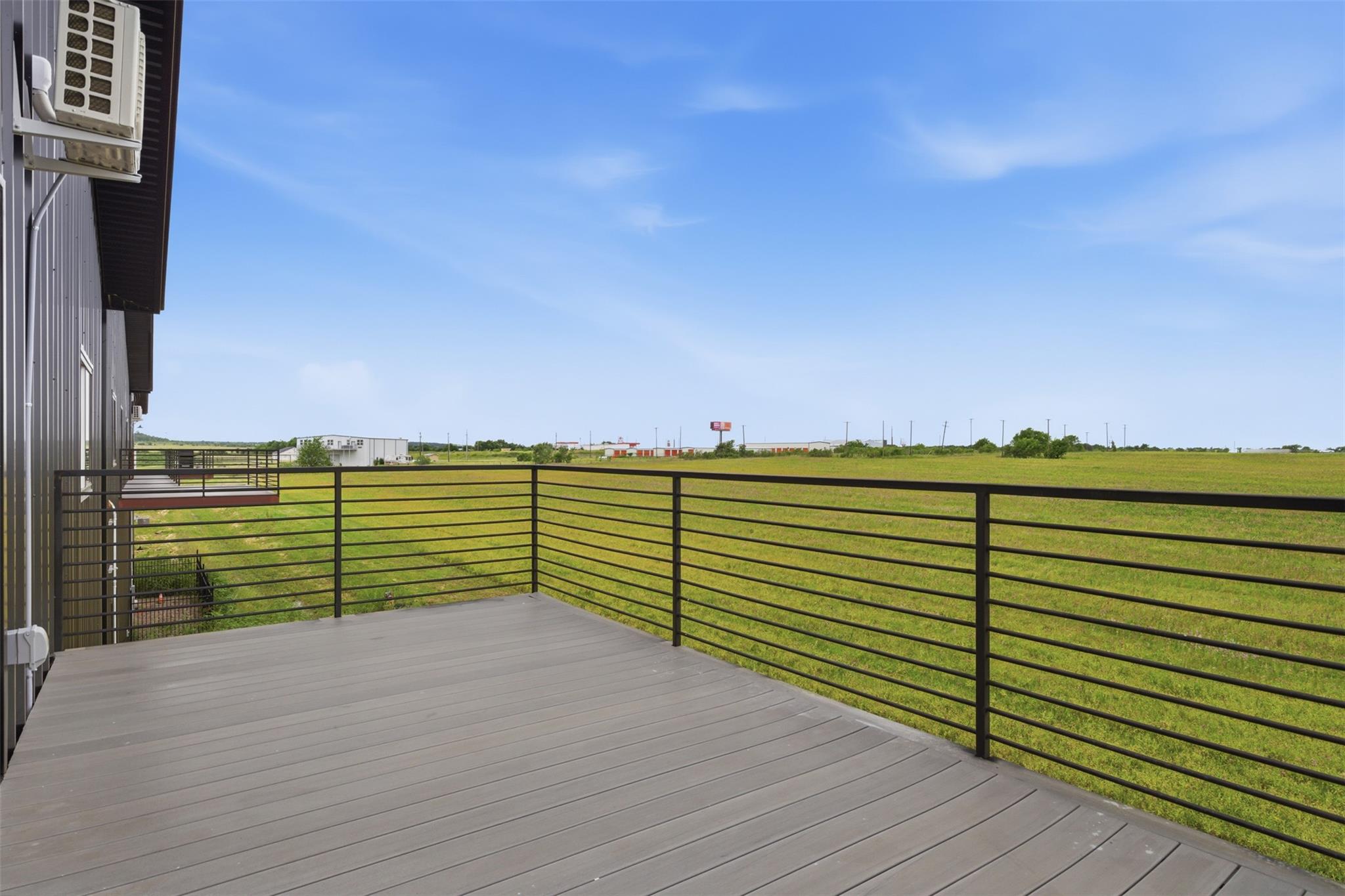 Gray deck with black railing beside a metal-sided building, overlooking a grassy field.