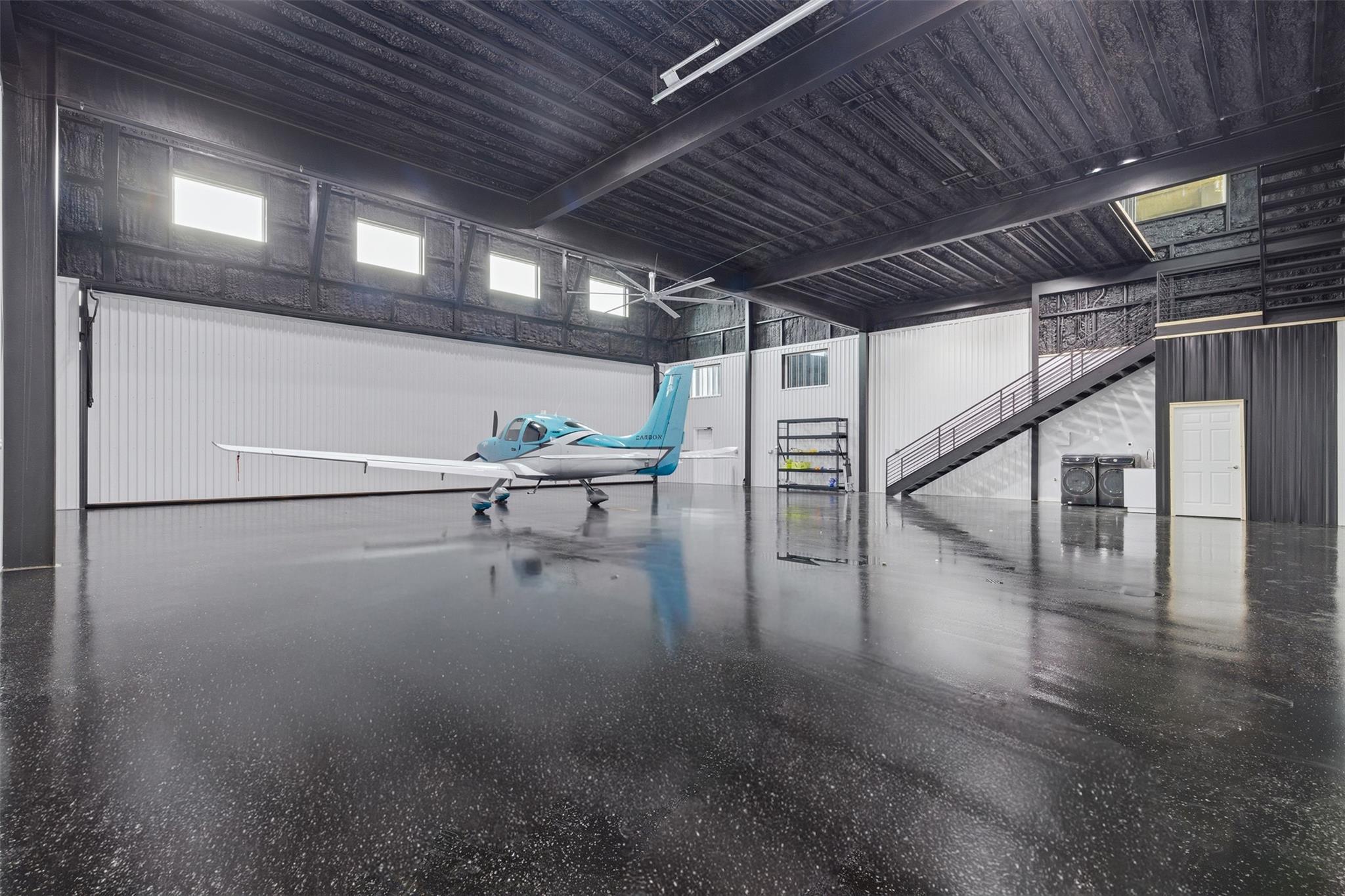 Aircraft hangar with a blue-and-white airplane, glossy floor, and stairs to a mezzanine.