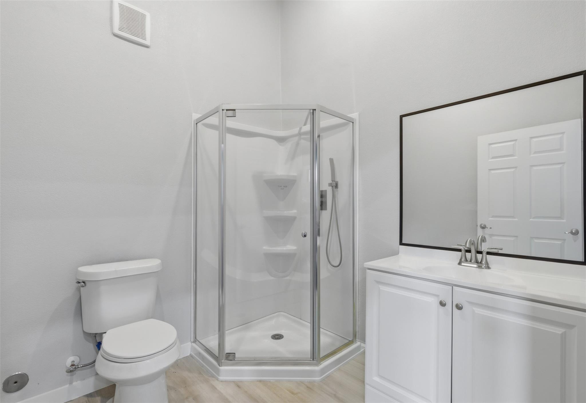 White bathroom with a corner glass shower, toilet, and vanity beneath a large mirror.