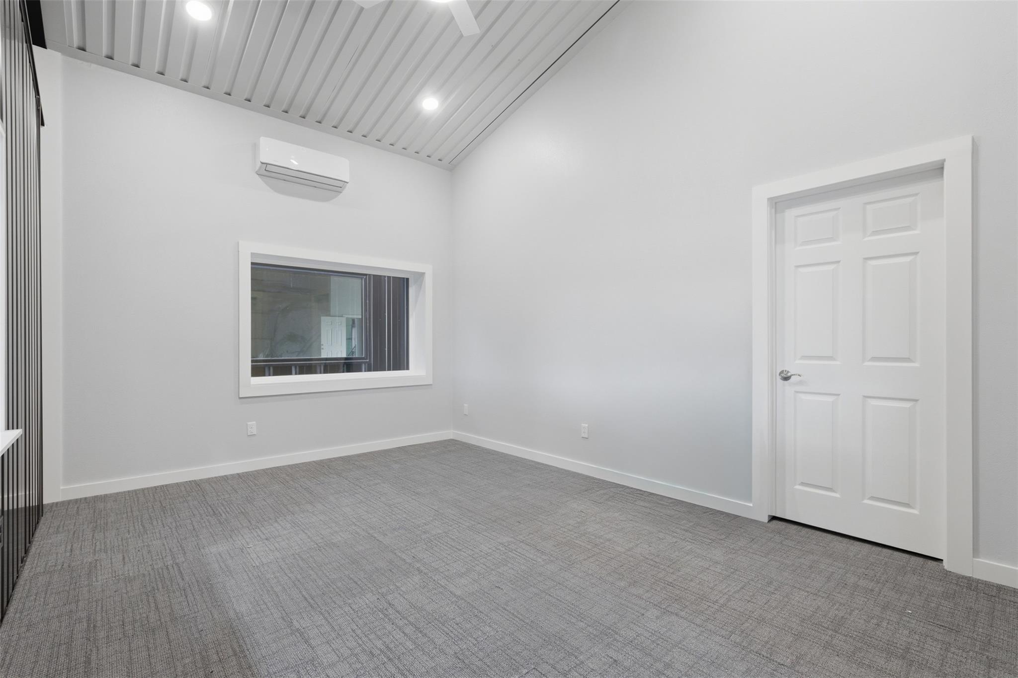 Empty carpeted room with a vaulted slatted ceiling, mini-split air conditioner, and interior window.