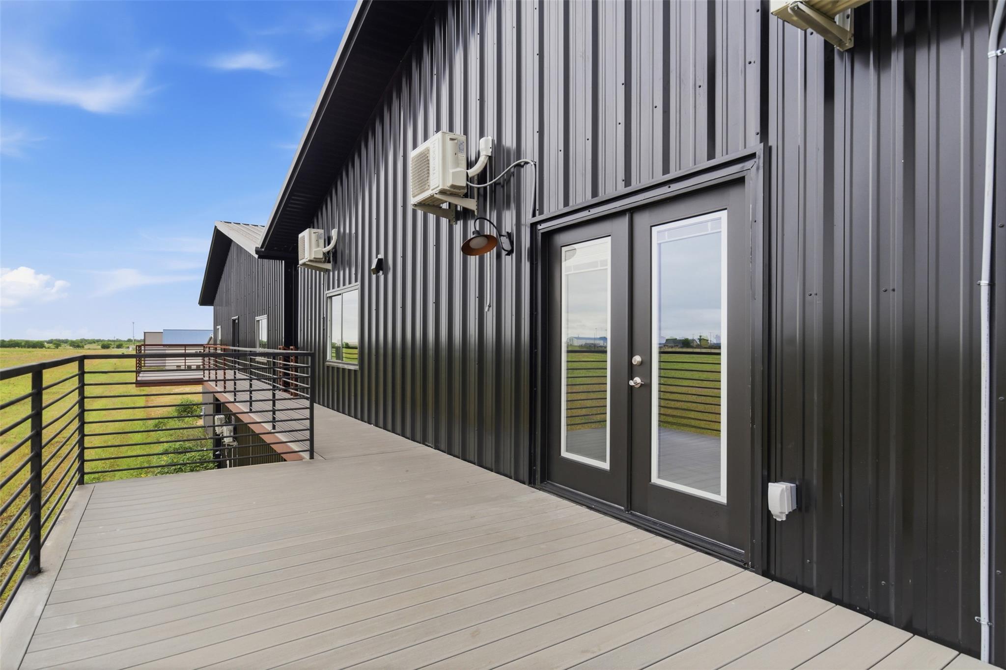 Wide gray deck along a black metal-sided building, with glass doors and railings overlooking grassy fields.