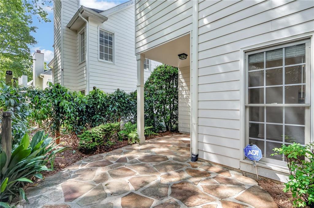 Stone patio beside a white-sided home, bordered by leafy shrubs and a covered walkway.