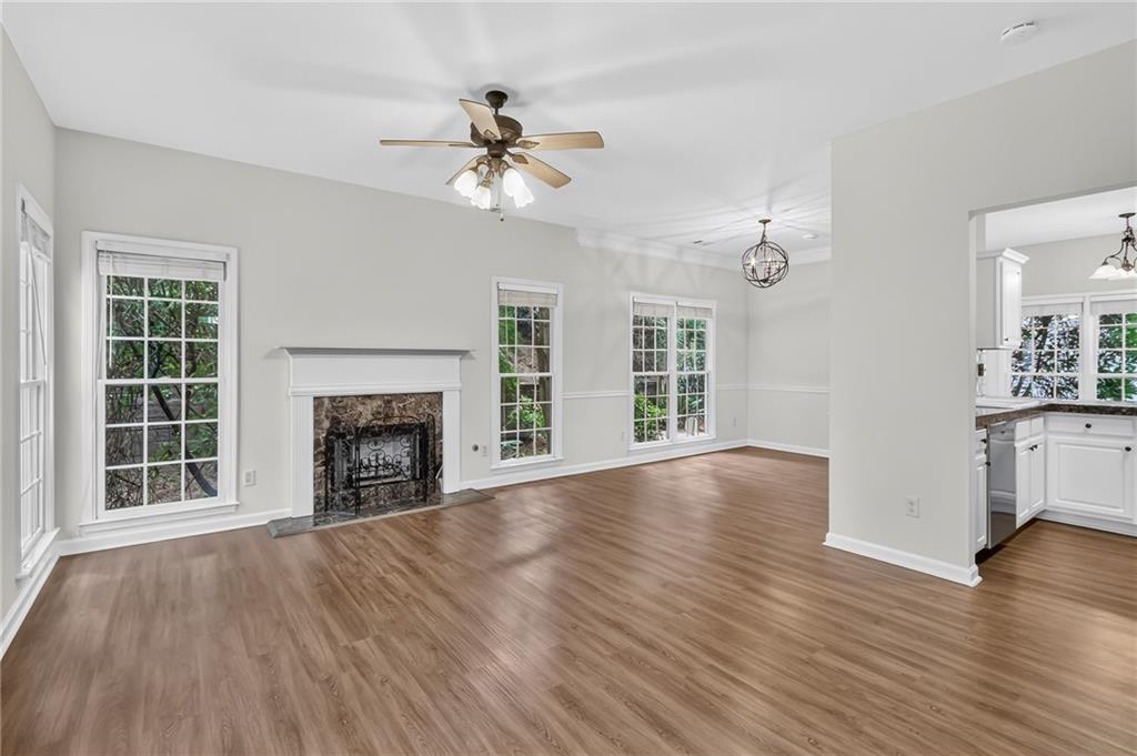 Vacant living room with wood flooring, a white-mantel fireplace, and windows beside an adjoining kitchen.