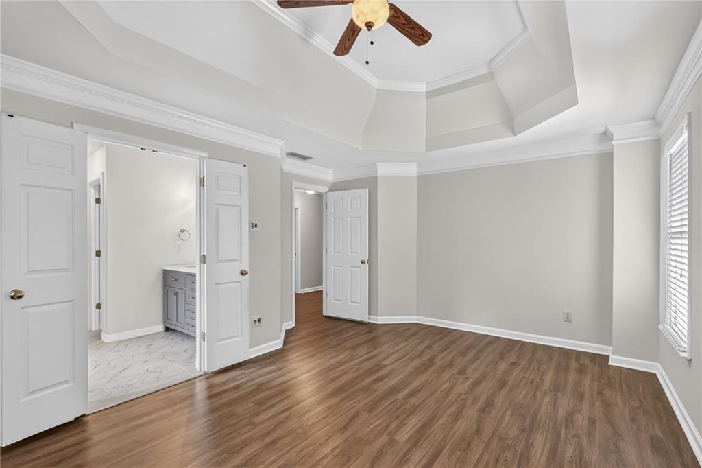 Empty bedroom with wood flooring, a tray ceiling, ceiling fan, and adjoining tiled bathroom.