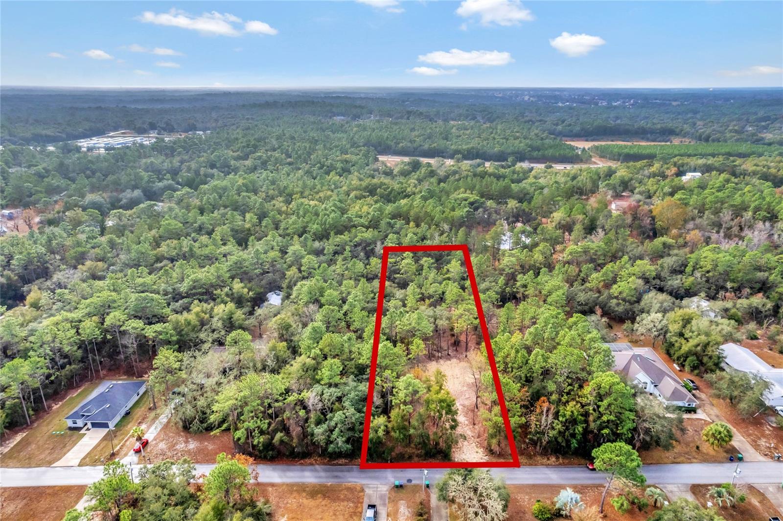 Aerial view of a wooded vacant lot outlined in red, with road frontage and a cleared area.