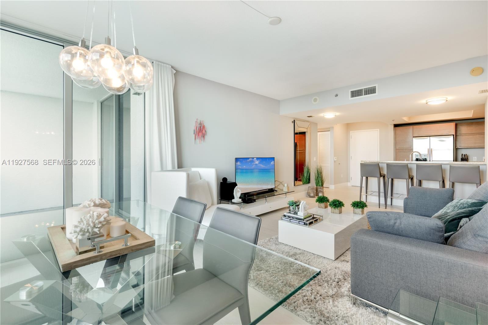 Open-plan condo living and dining area with a glass table, gray sectional, and kitchen island.