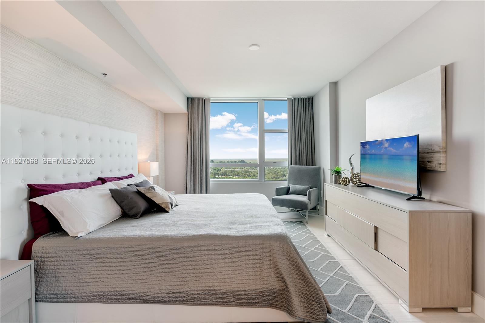 Furnished bedroom with a tufted white headboard, gray chair, and broad windows overlooking greenery.