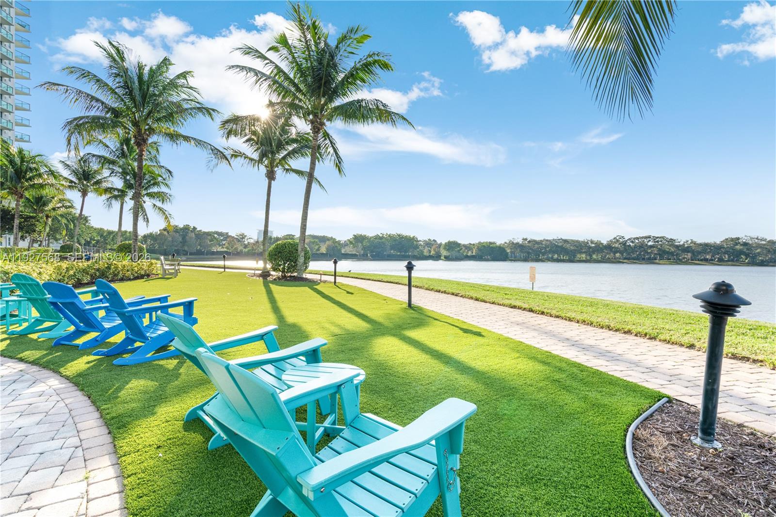 Lakeside lawn with turquoise Adirondack chairs, palm trees, and a paved waterside path.