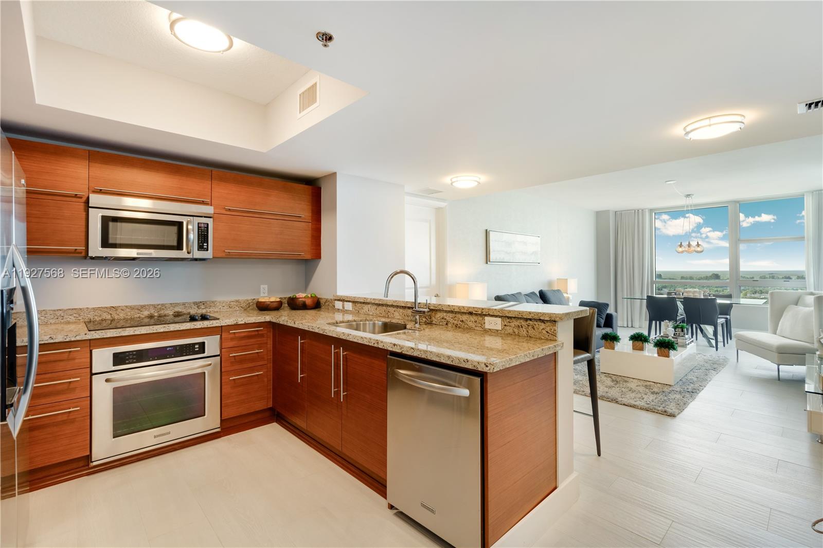 Modern wood-cabinet kitchen opening to a furnished living and dining area with a distant water view.