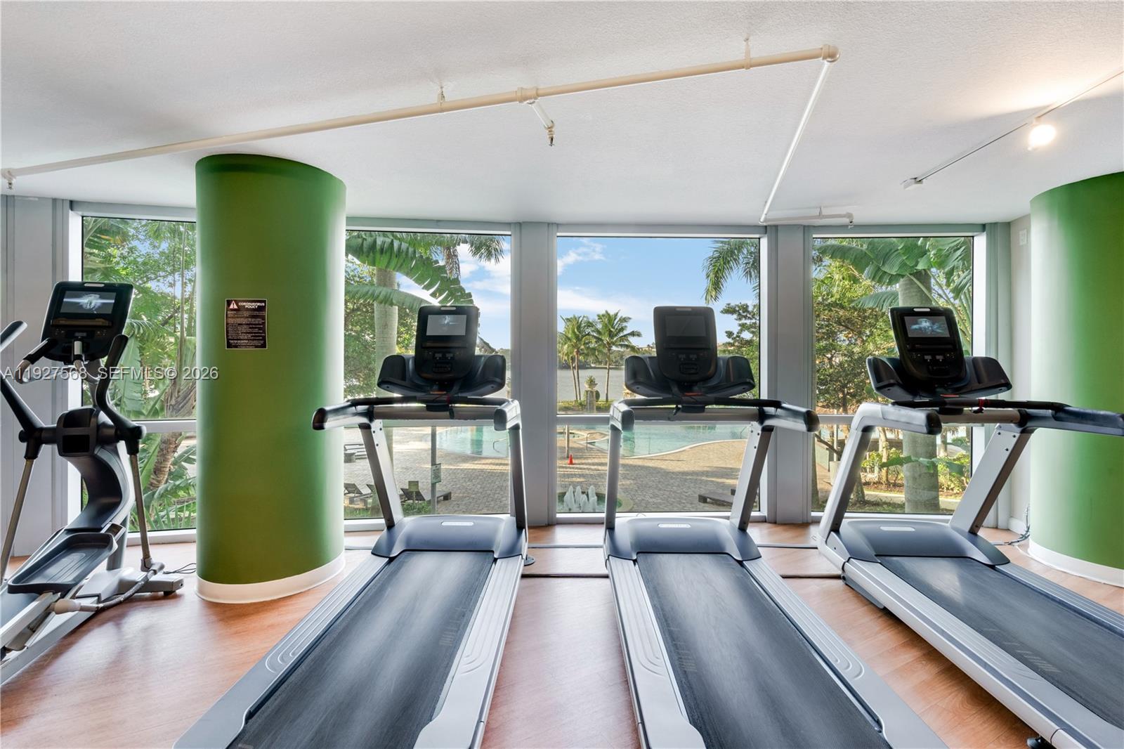 Condo gym with treadmills and an elliptical overlooking a pool and water through large windows.