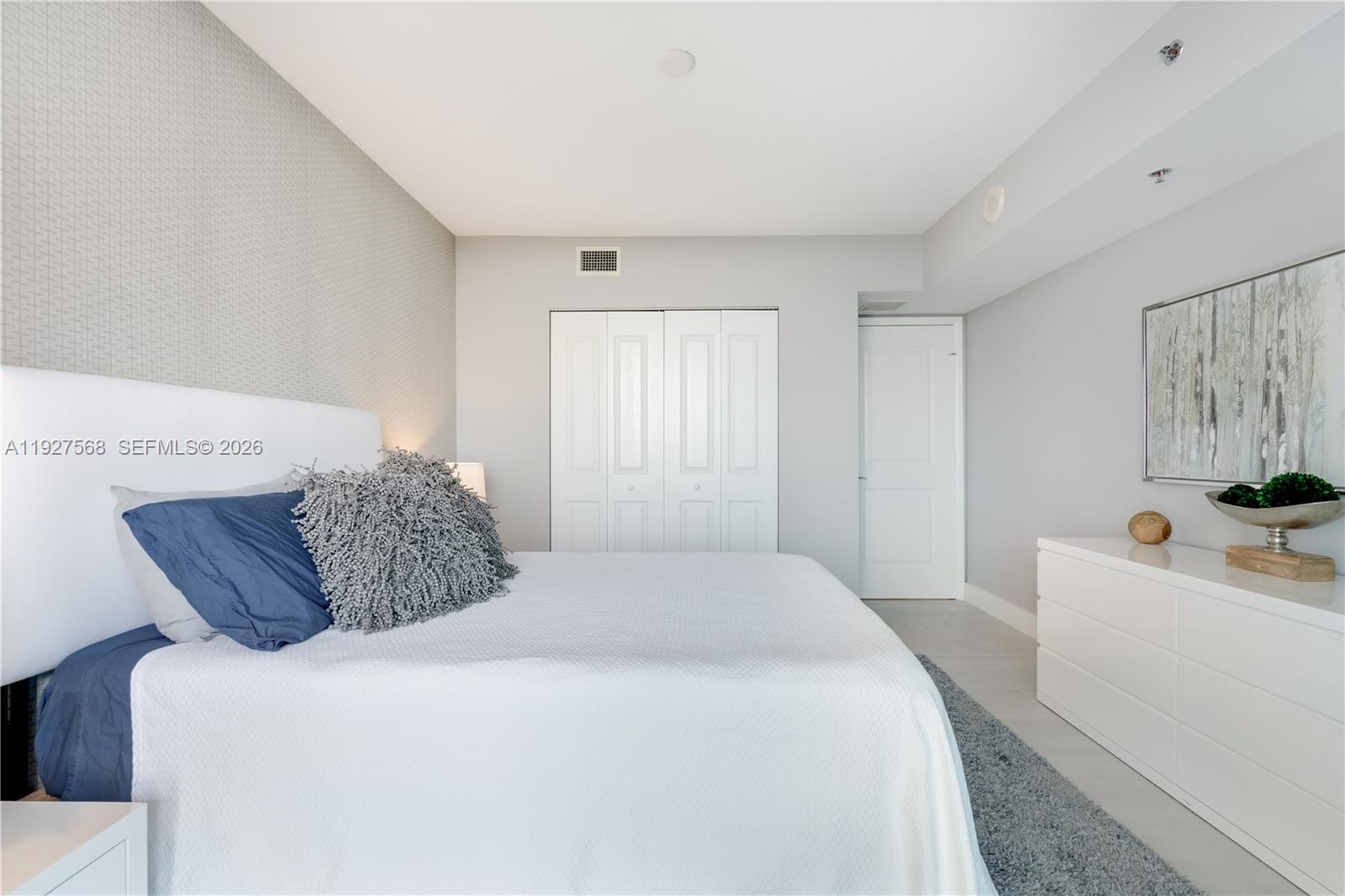 Staged bedroom with white bed, navy and gray pillows, patterned accent wall, and long white dresser.
