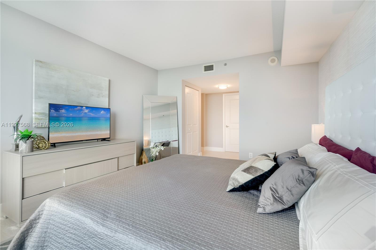 Bedroom with a gray quilted bed, tufted headboard, and television displaying a beach scene.