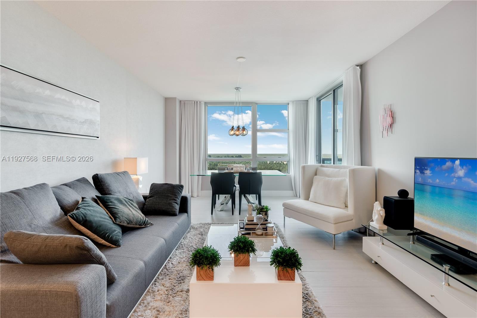 Furnished condo living room with a gray sectional, white chair, and large windows overlooking greenery.
