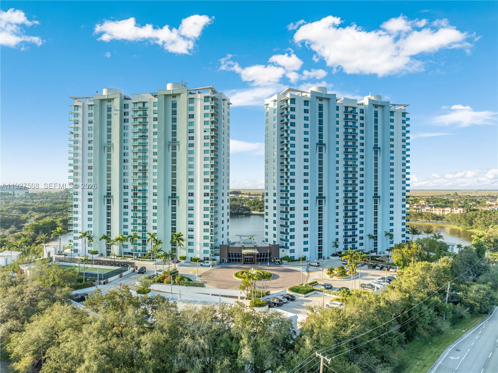 Two white condominium towers with turquoise balconies stand beside a waterway amid palm trees and greenery.