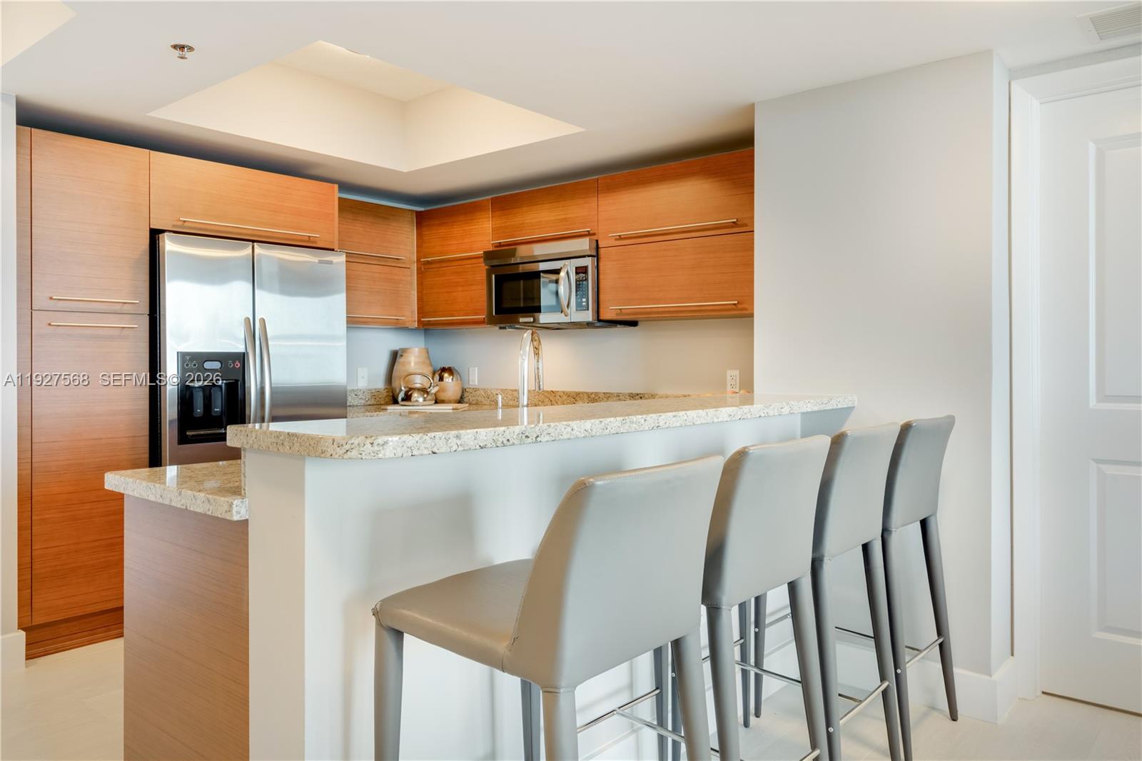 Modern kitchen with warm wood cabinets, stainless steel appliances, and a stone breakfast bar with gray stools.