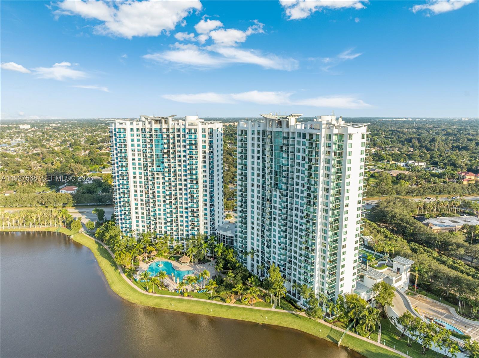 Two white condominium towers beside a lake, with a landscaped pool area and palm trees.