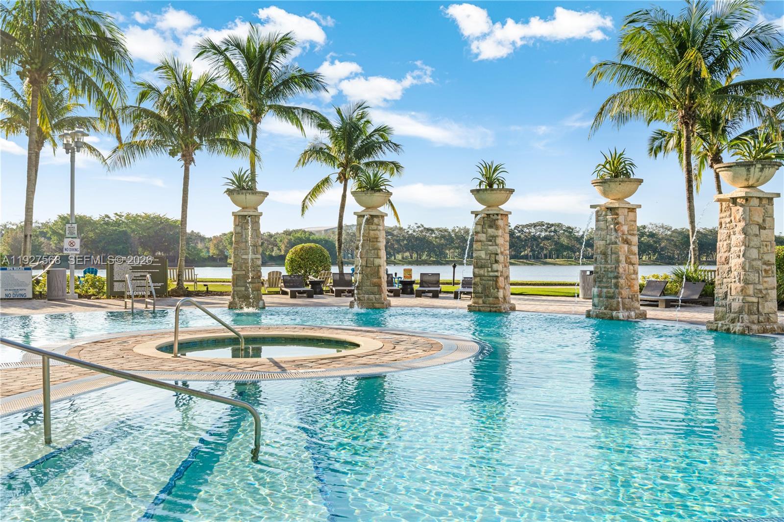 Outdoor pool and circular spa with palm trees, stone fountain columns, and a lake beyond.