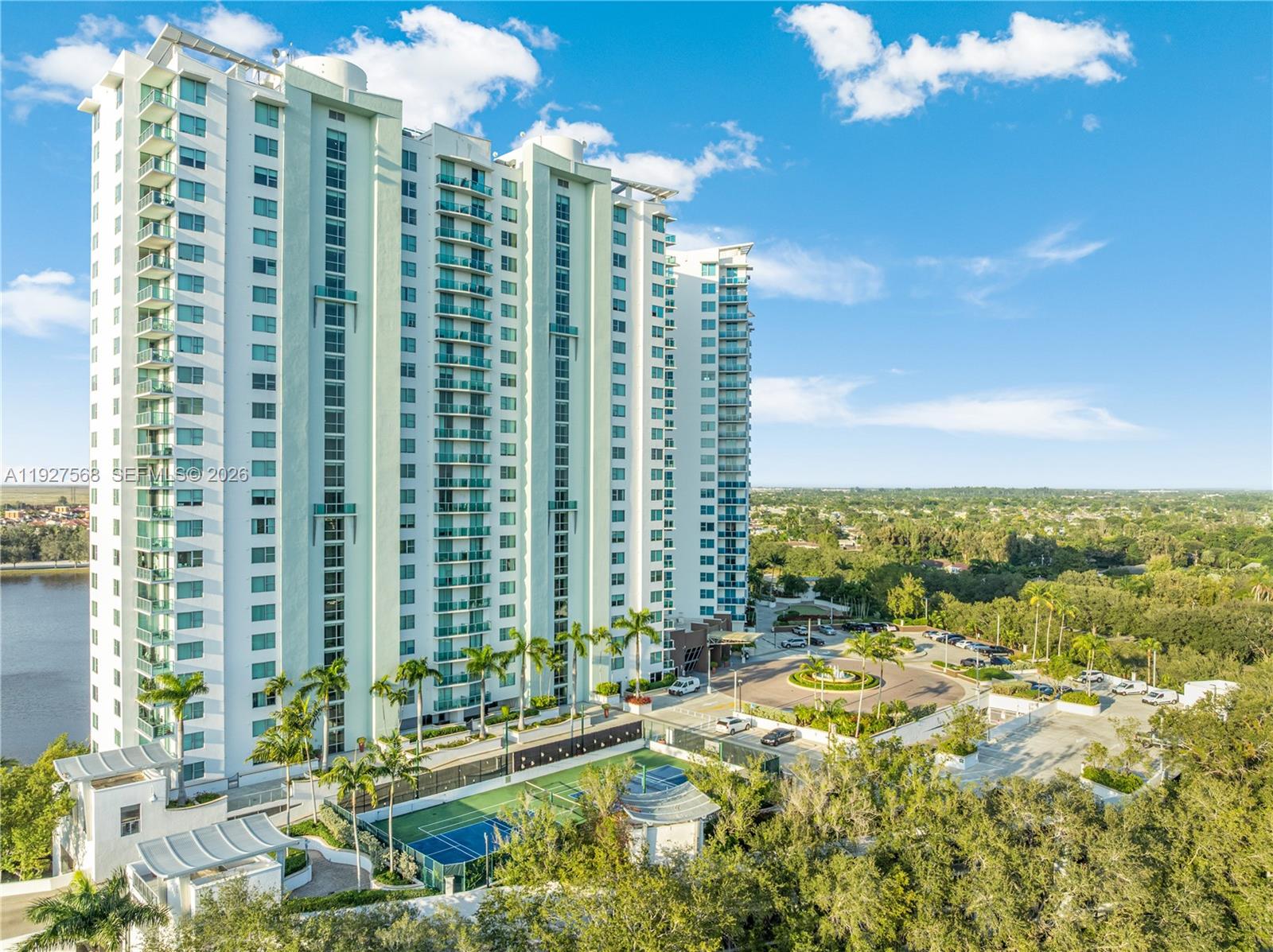 White high-rise condominium with glass balconies, landscaped grounds, and a tennis court beside a lake.