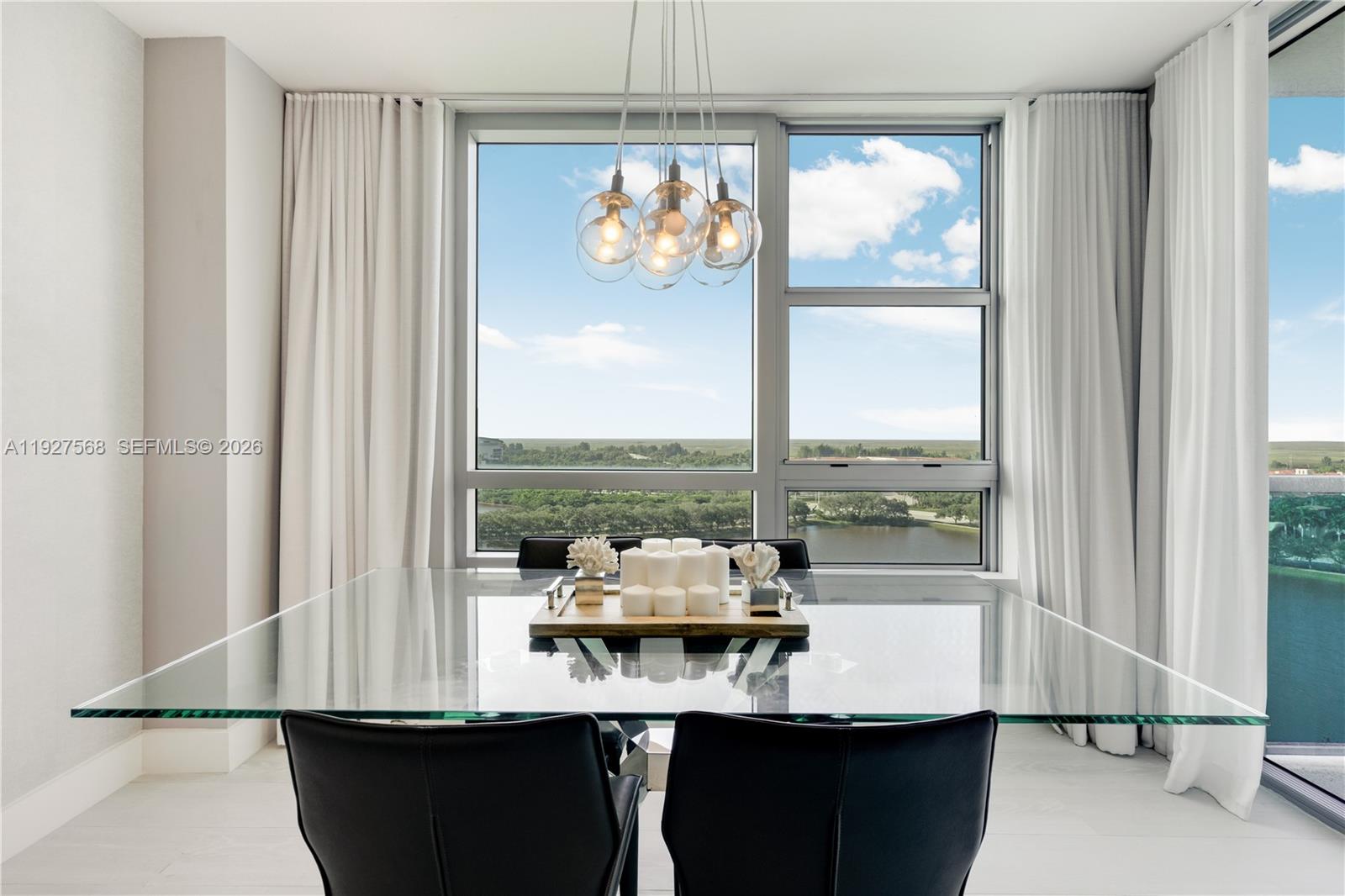 Dining area with a glass table and black chairs facing floor-to-ceiling windows overlooking a lake.
