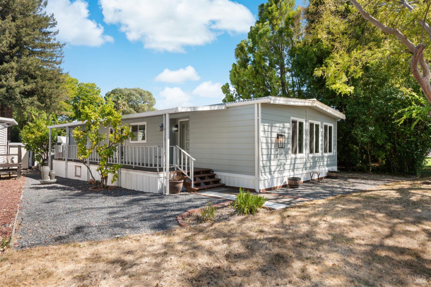 Light gray manufactured home with a covered porch, gravel yard, and mature trees.