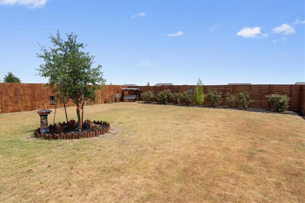 Fenced backyard with a broad lawn, a mature tree in a circular planting bed, and shrubs along the fence.