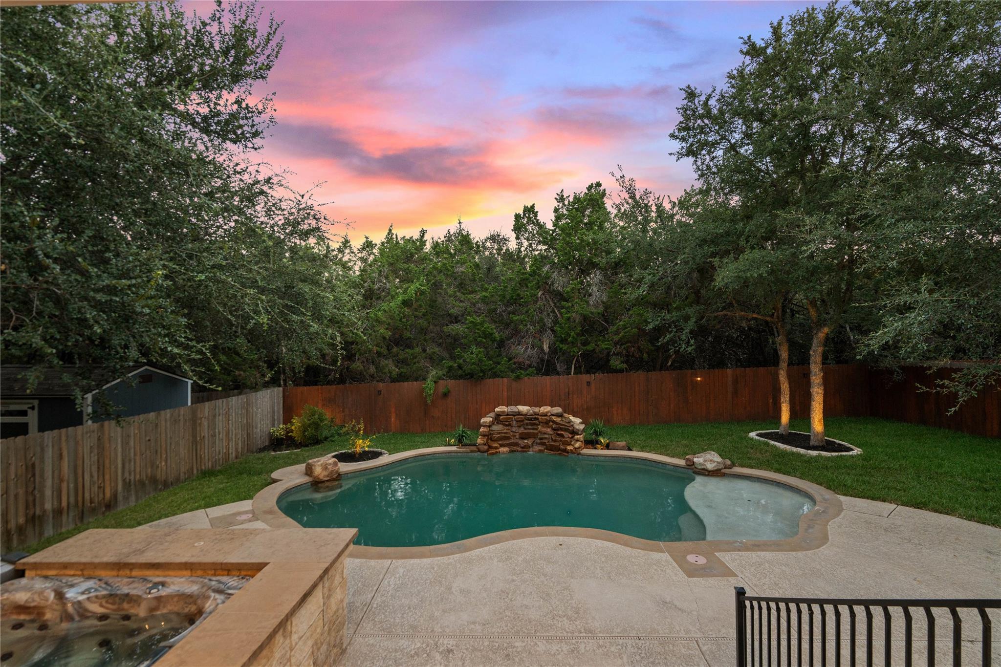 Backyard pool and stone spa beside a fenced lawn at sunset.