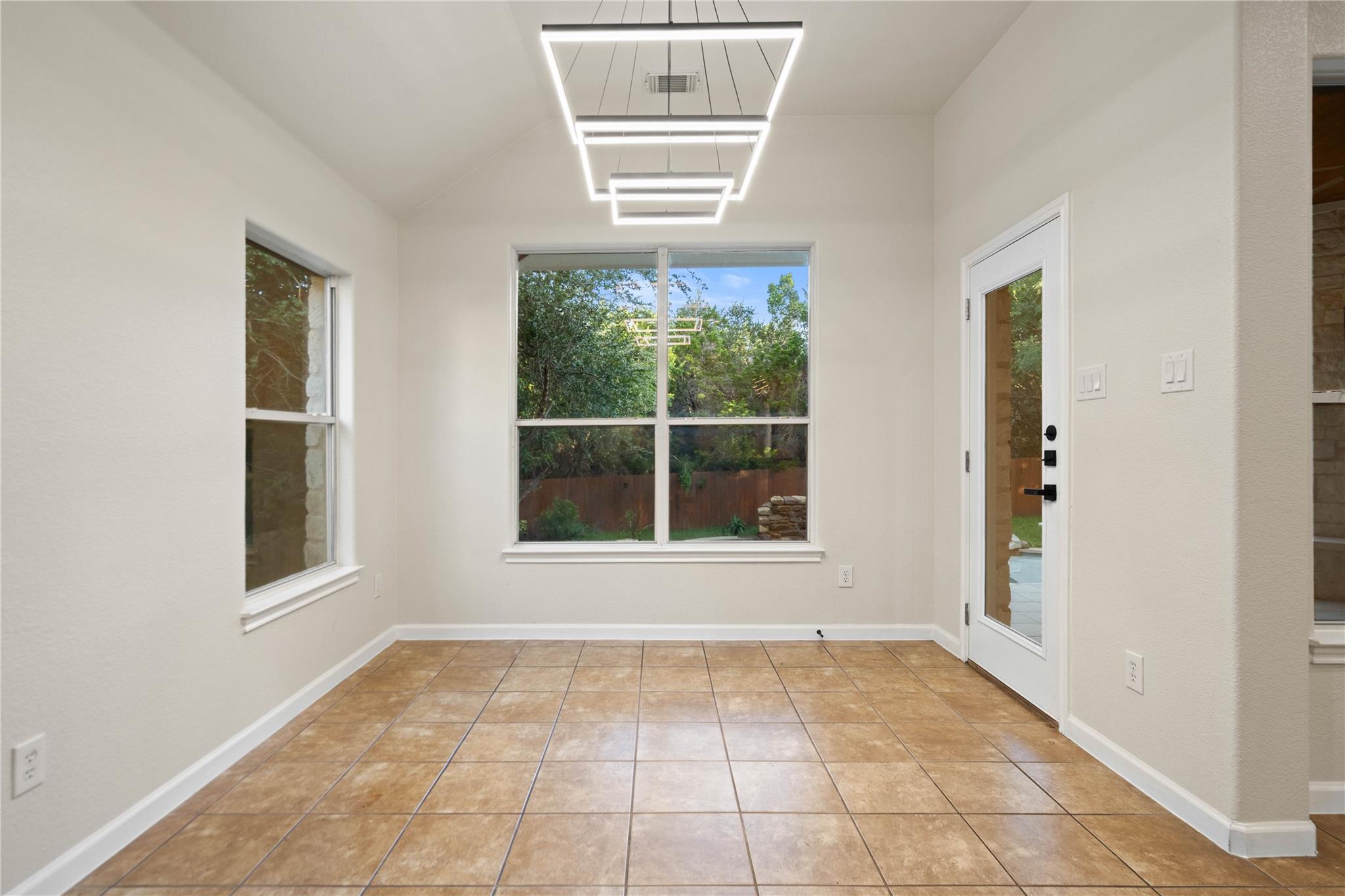 Bright sunroom with tan tile flooring, geometric pendant lights, and windows overlooking a fenced yard.
