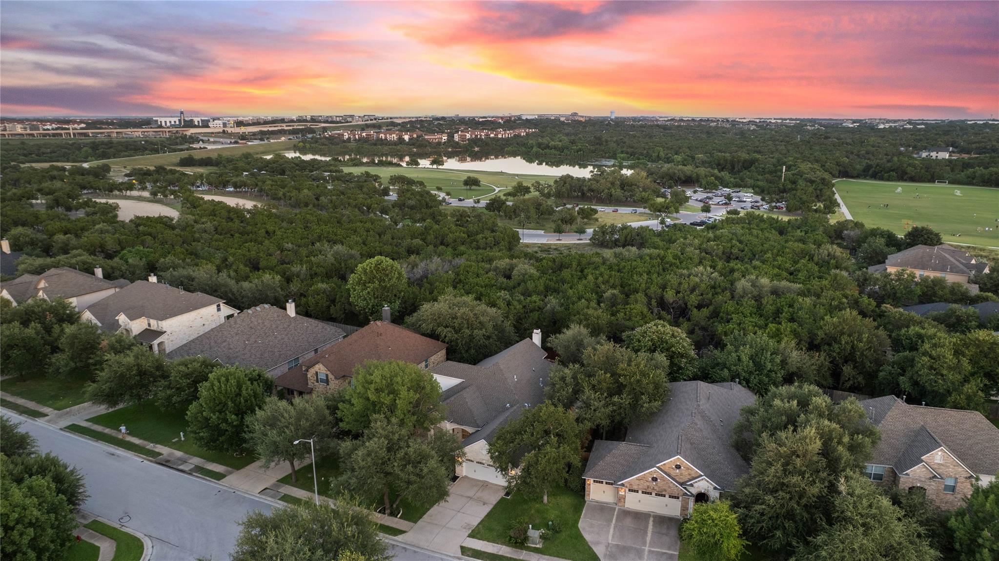 Aerial view of homes beside a wooded park, pond, and sports fields at sunset.