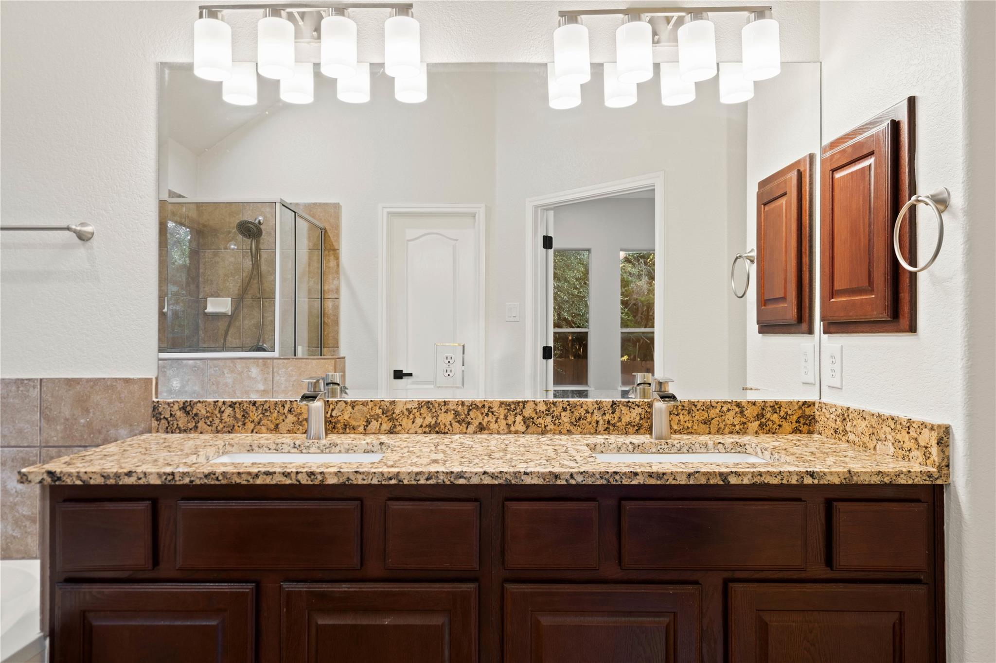Double-sink bathroom vanity with granite counters, dark wood cabinets, and a tiled shower.