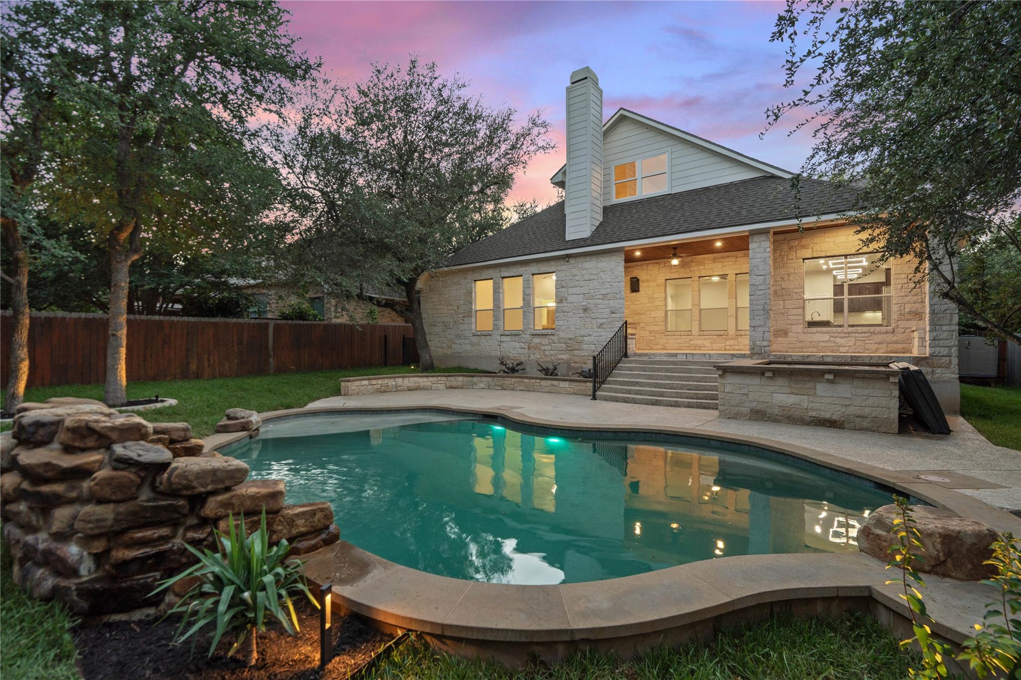 Stone house overlooks an illuminated backyard pool at dusk.