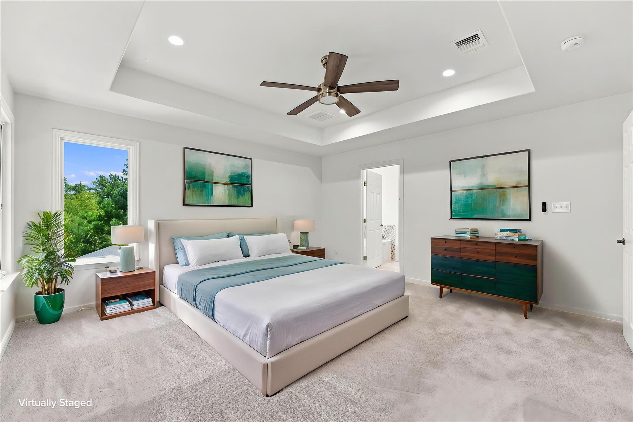 Virtually staged bedroom with a large bed, tray ceiling, dark wood fan, and a window overlooking trees.