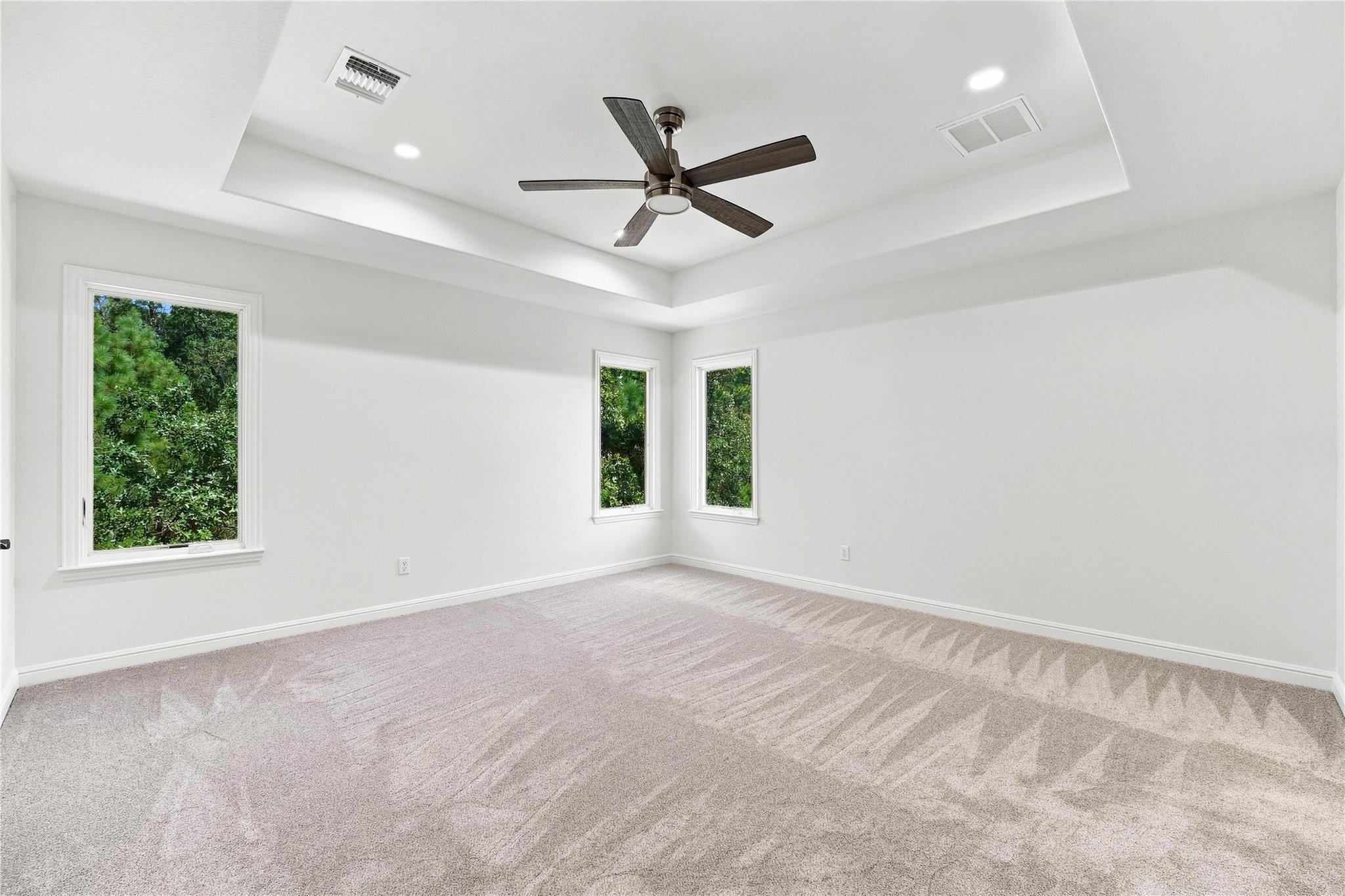 Empty carpeted room with a tray ceiling, dark ceiling fan, and windows overlooking greenery.