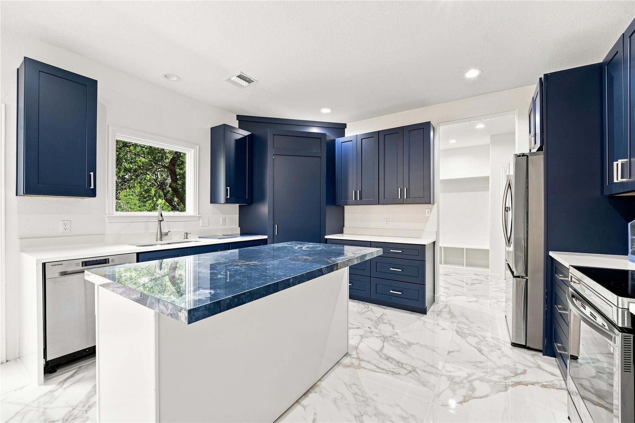 Modern kitchen with navy cabinets, blue-stone island, stainless appliances, and marble-look tile floors.
