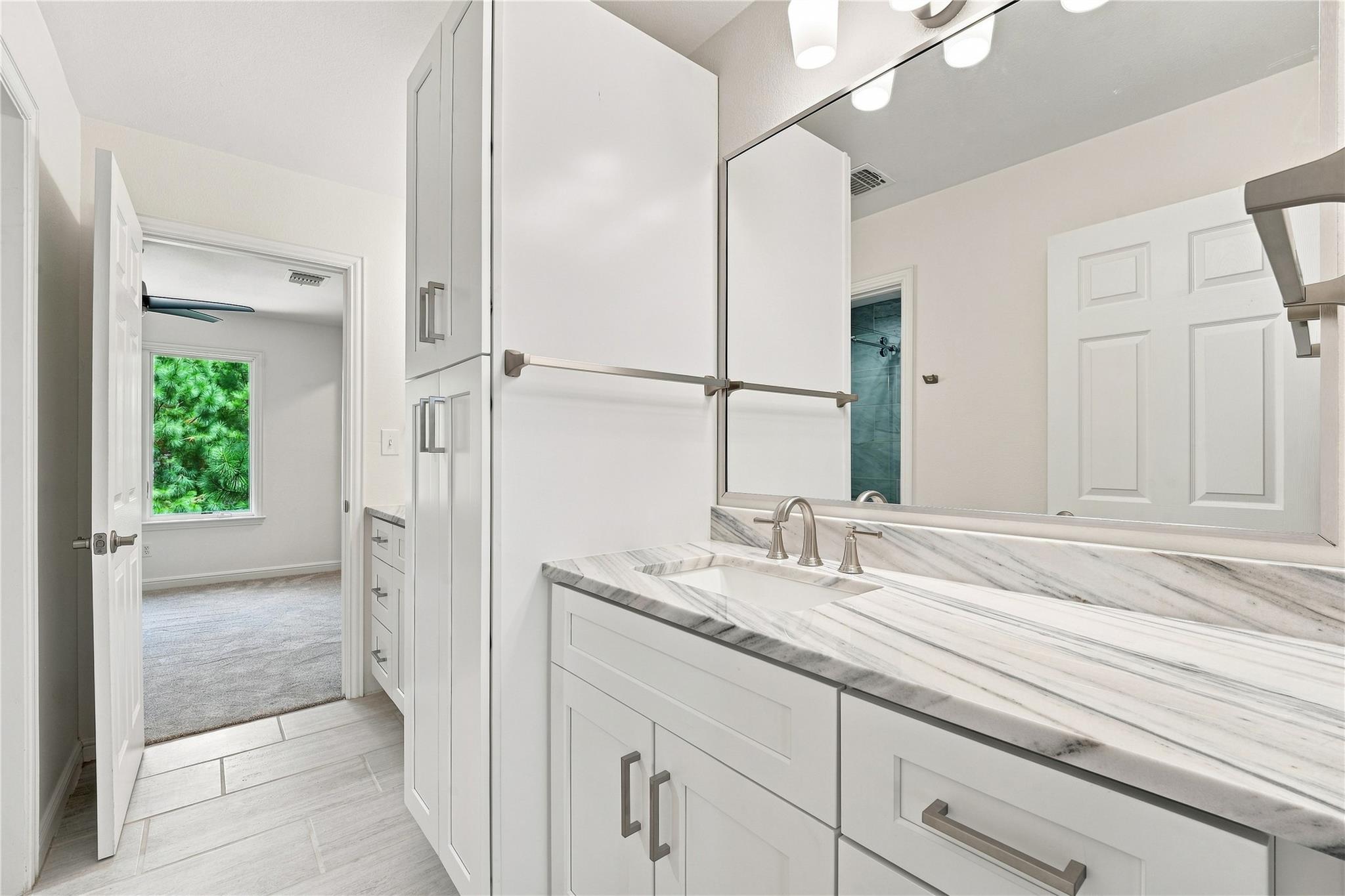 Bathroom vanity with a gray-veined stone counter and large mirror beside an open doorway to a carpeted room.