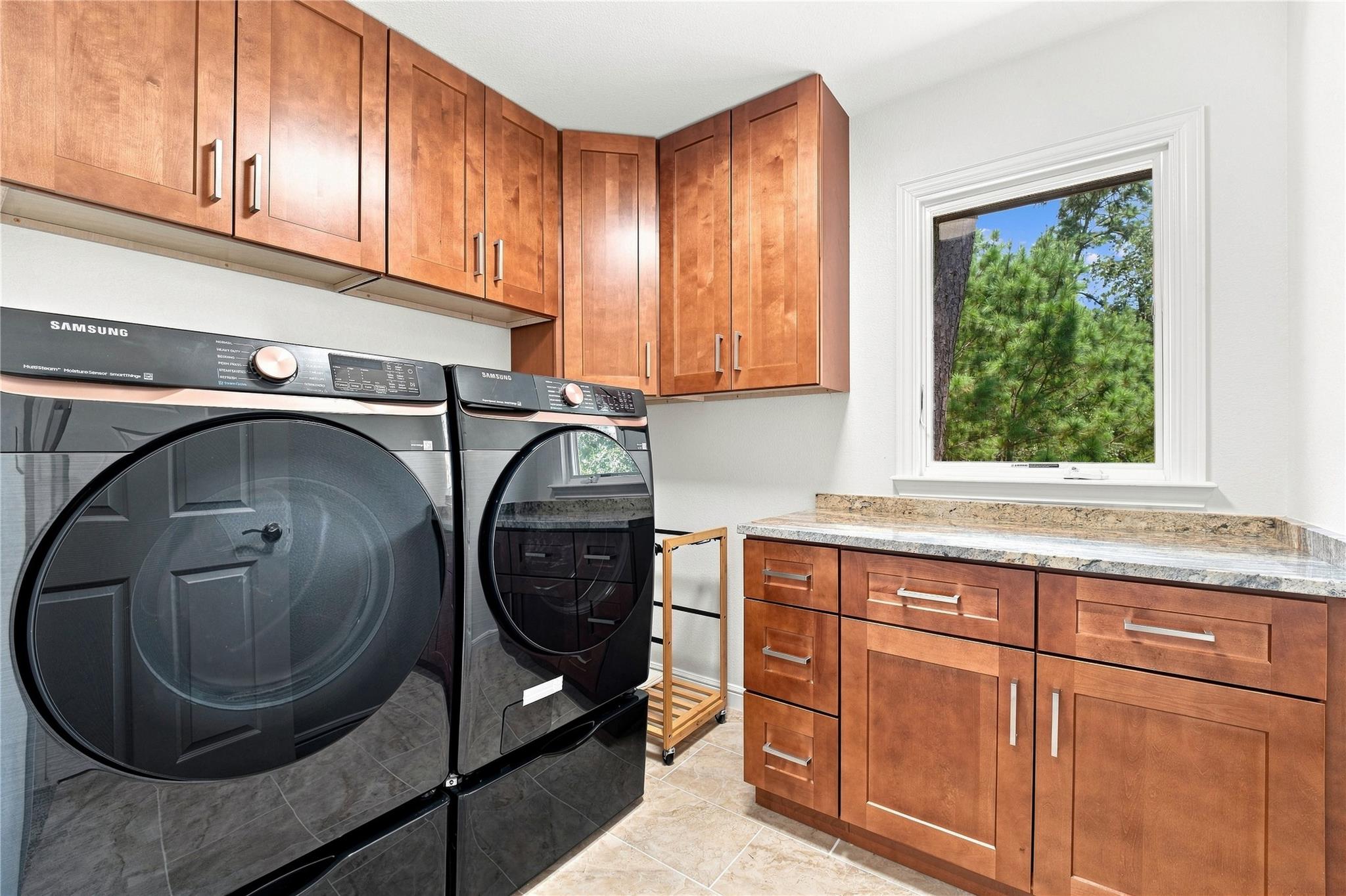 Laundry room with Samsung washer and dryer, warm wood cabinetry, stone counter, and a window overlooking trees.