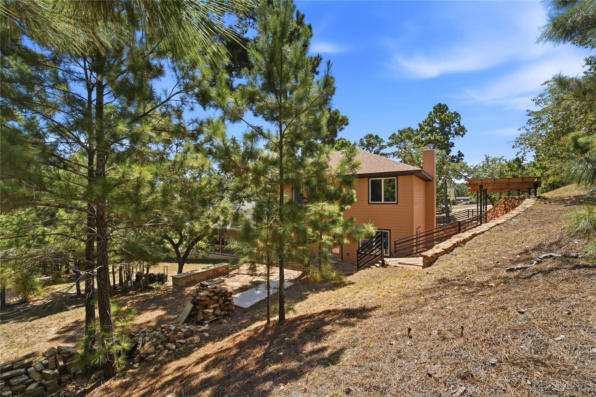 Tan two-story home among pine trees, with a sloped yard, stone walls, and a wooden pergola.